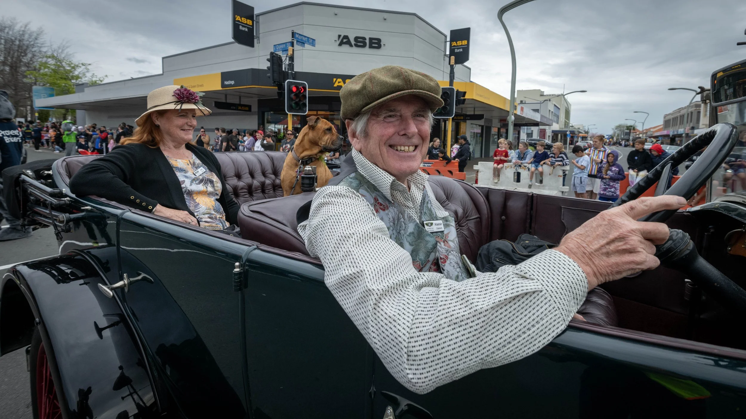 People riding in a vintage car during the Hastings blossom parade, with a crowd of spectators in the background on a city street.