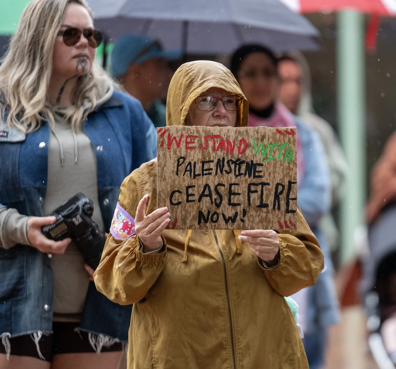 A woman wearing a yellow raincoat and glasses holding a cardboard protest sign that reads 'WE STAND WITH PALENSTINE CEASE FIRE NOW!' during a rainy outdoor rally, with other people and umbrellas in the background.