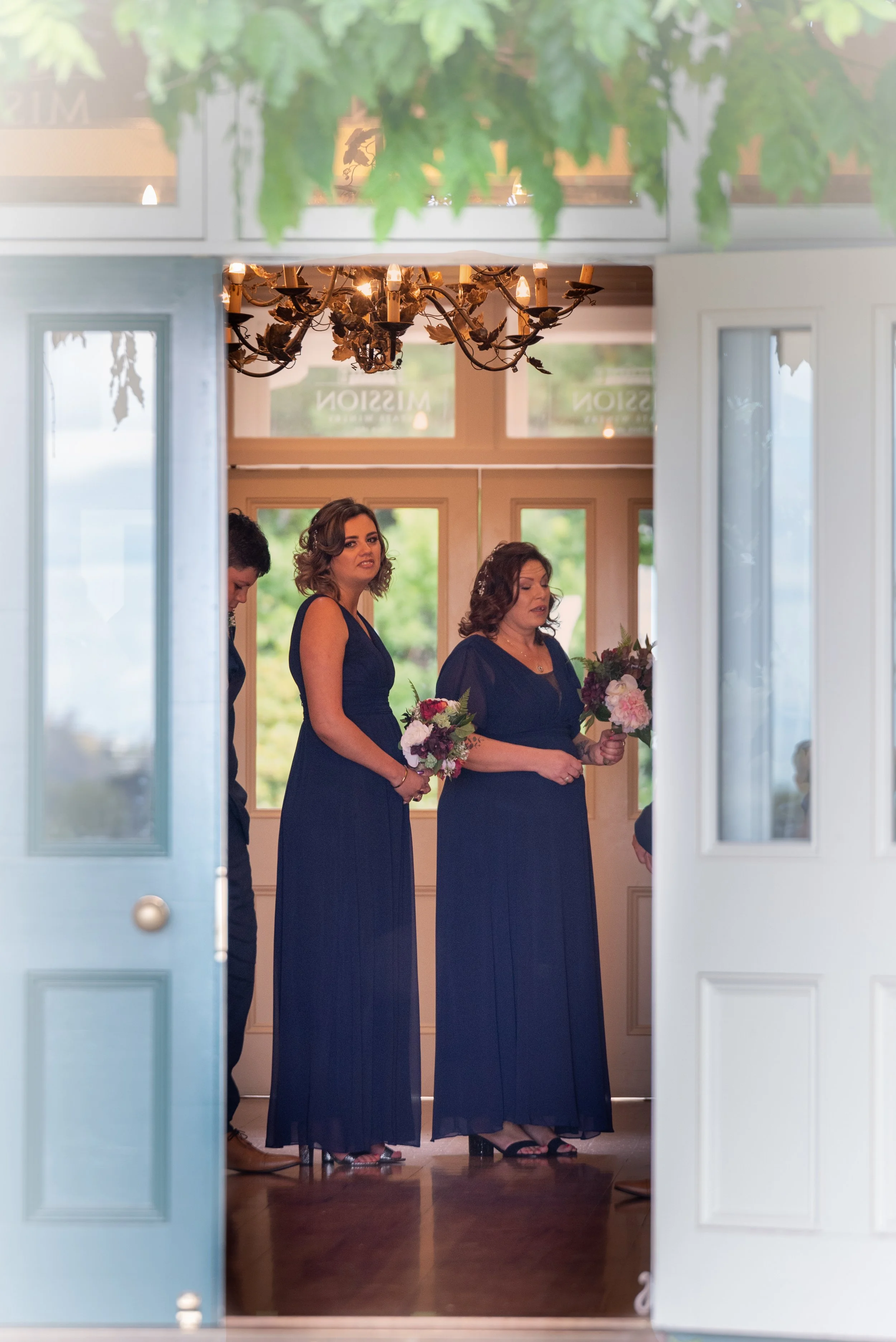 Three women in navy blue dresses holding bouquets, standing inside a building with wooden floors, a chandelier, and large windows, viewed through an open door.