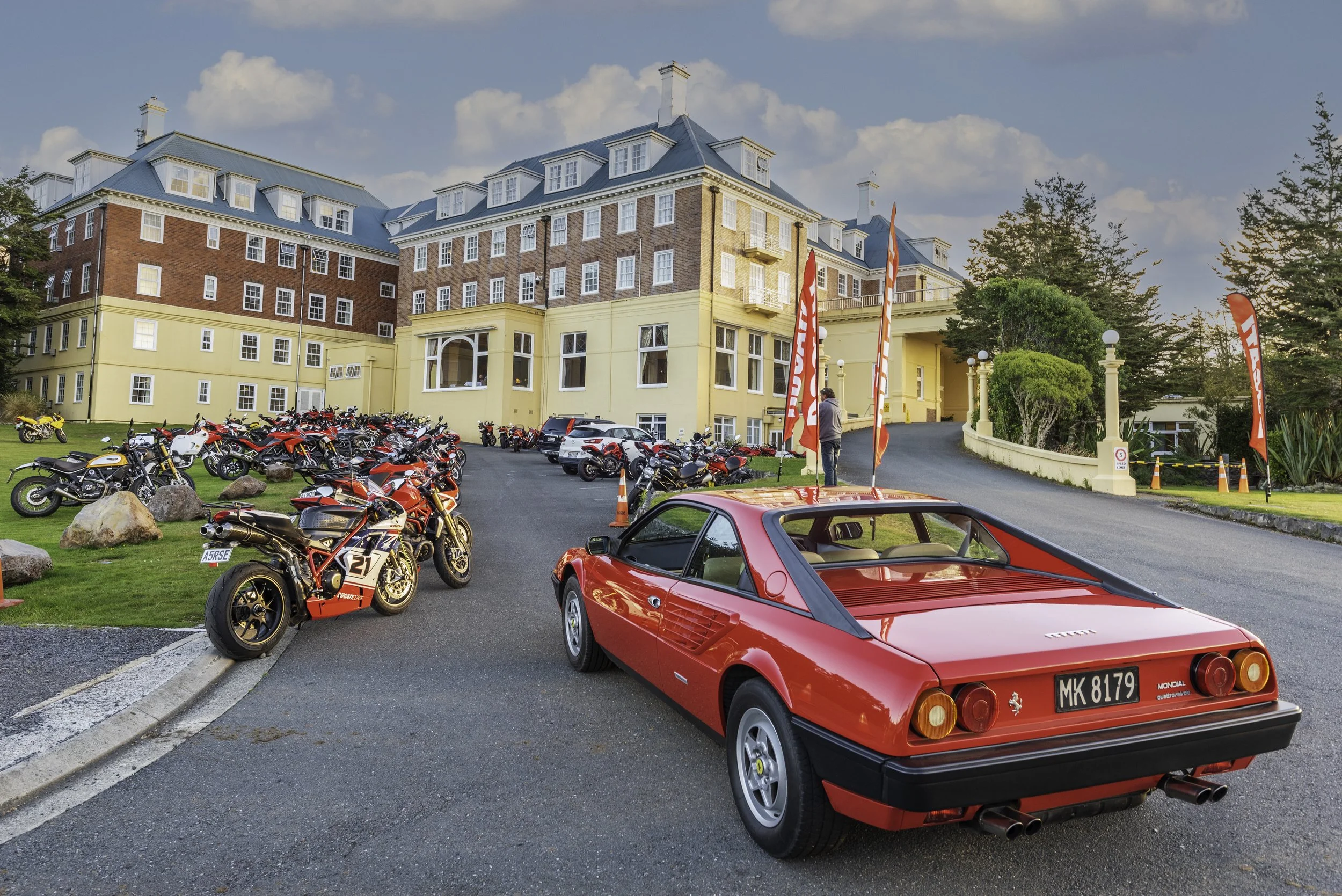 A parking lot in front of a large, multi-story building with a blue roof and brick walls. The lot is filled with various motorcycles and a few cars, including a red sports car in the foreground and a Ferrari with a license plate. Flags and trees are 