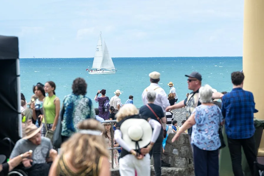Crowd of people gathered on a seaside promenade with sailboats in the ocean.