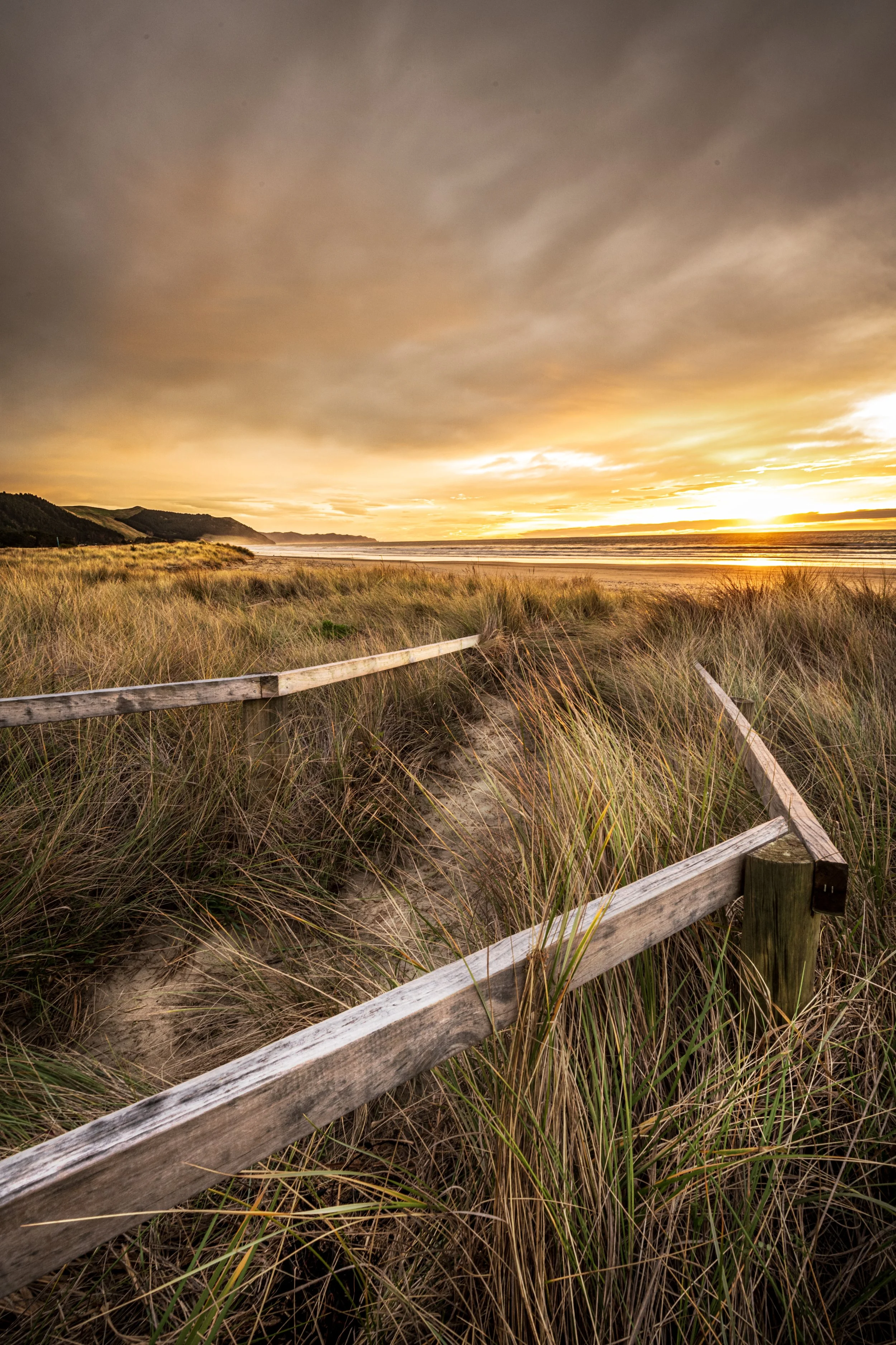 Sunset over a beach with grassy dunes and a wooden fence in the foreground.