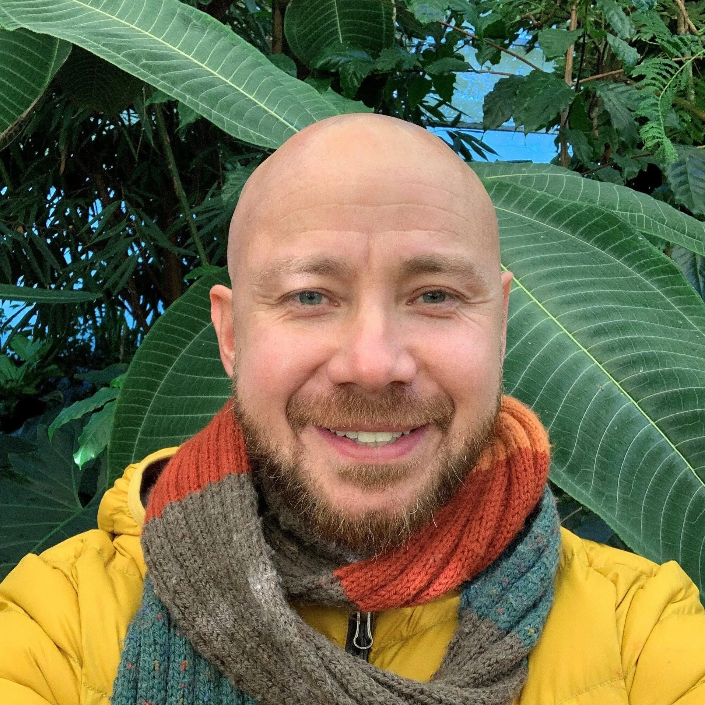A person with blue eyes and a light beard smiling, wearing a yellow jacket, a multicolored scarf, and standing in front of large green tropical leaves.