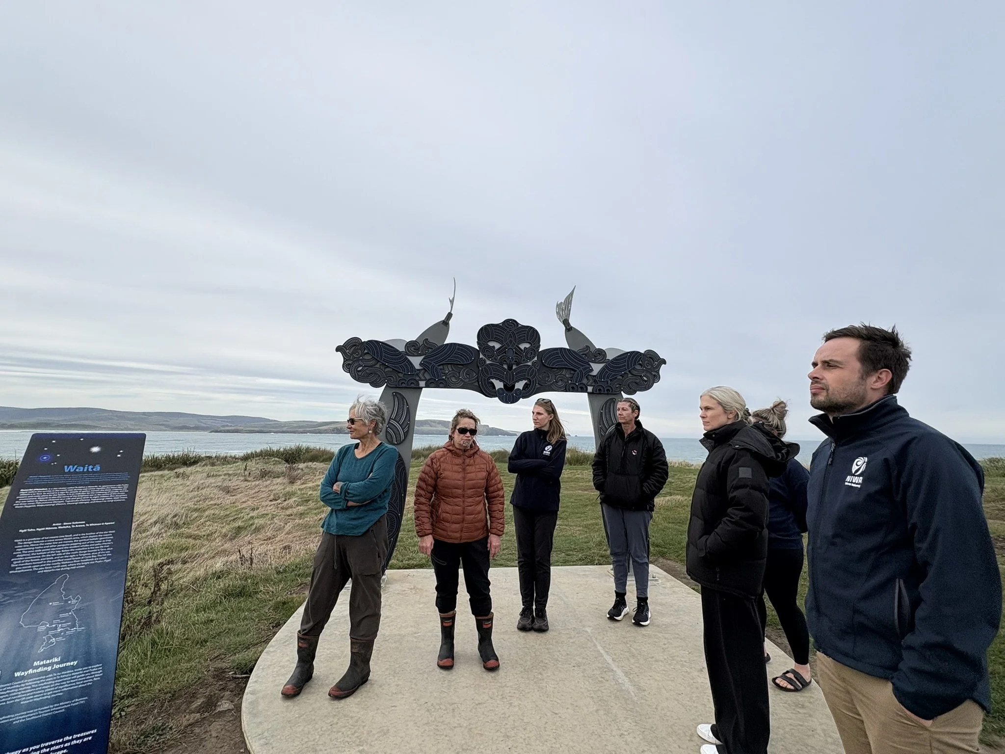A group of people listening to a speaker outdoors near a large sign and an artistic sculpture, with a body of water and hills in the background.