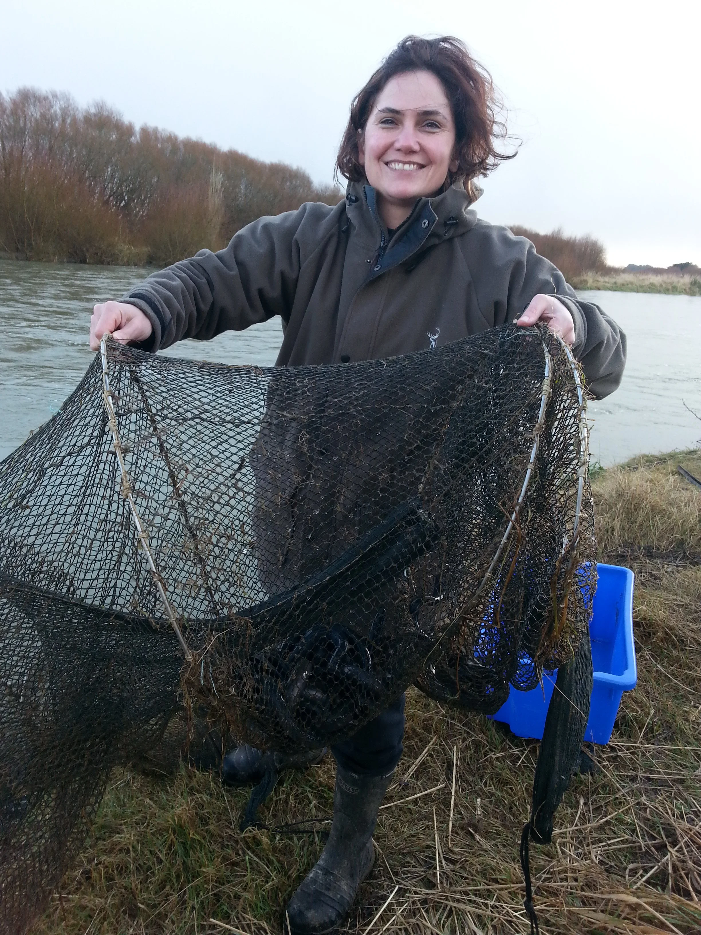 A woman in outdoor clothing smiling and holding a fishing net near a river, with trees in the background on a cloudy day.