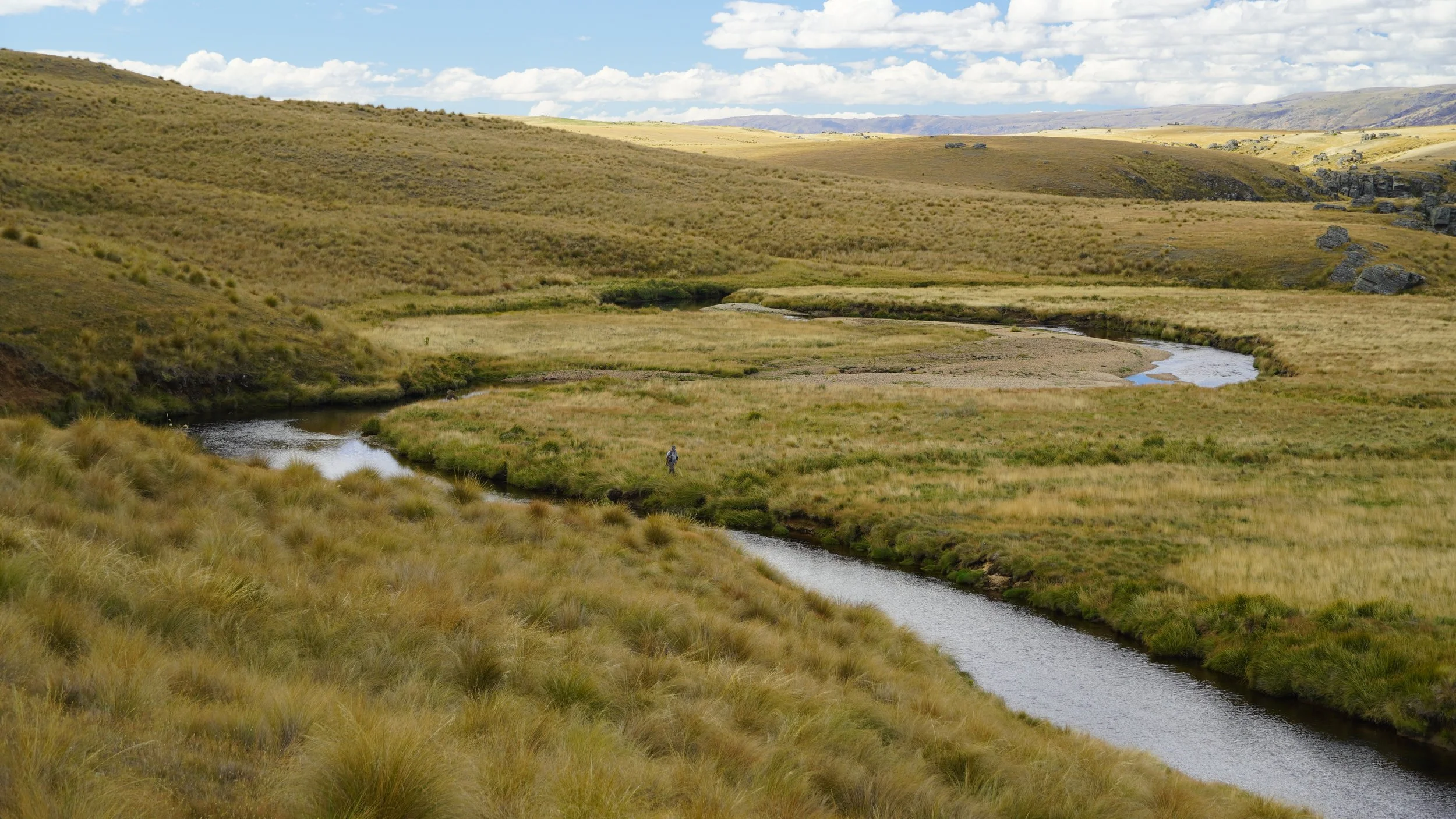 A vast landscape with rolling grassy hills, a winding river, and scattered rocks under a partly cloudy sky. A lone person is walking near the river.