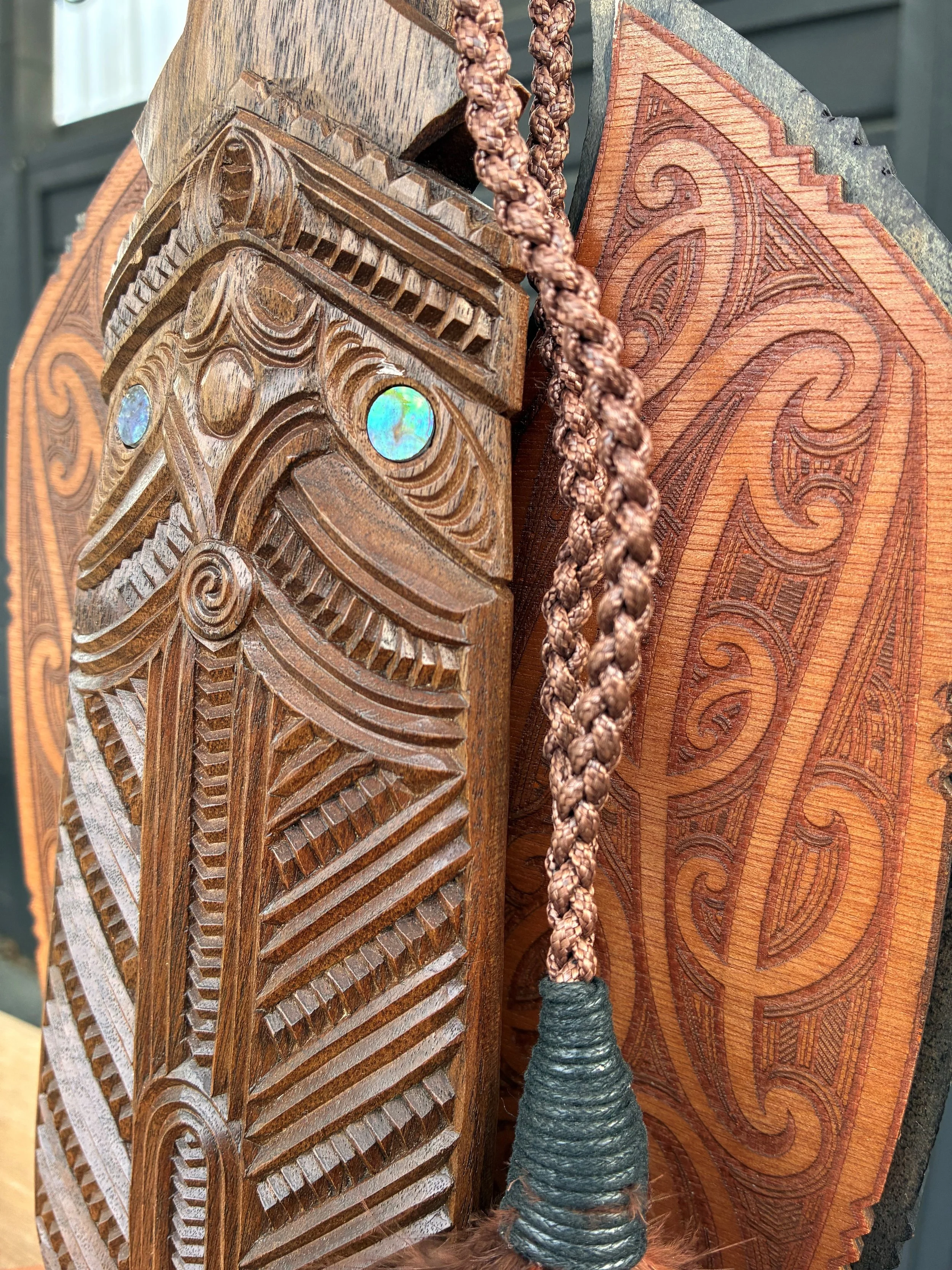 Close-up of carved wood award featuring intricate designs and face, with a braided cord hanging beside it, and another wooden surface with engraved patterns in the background.