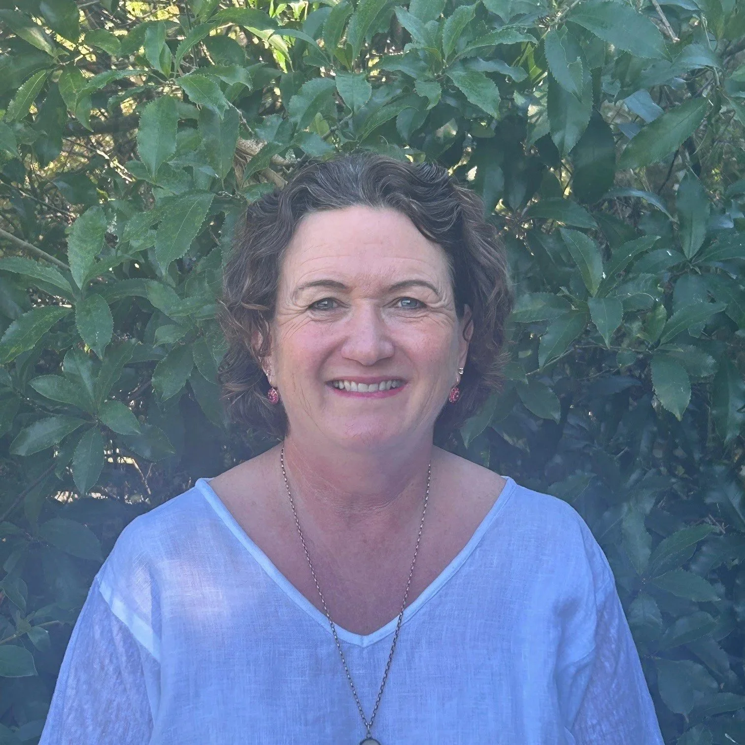 A smiling woman with short, curly hair standing outdoors in front of lush green leaves. She is wearing a light blue top, a necklace, and earrings.