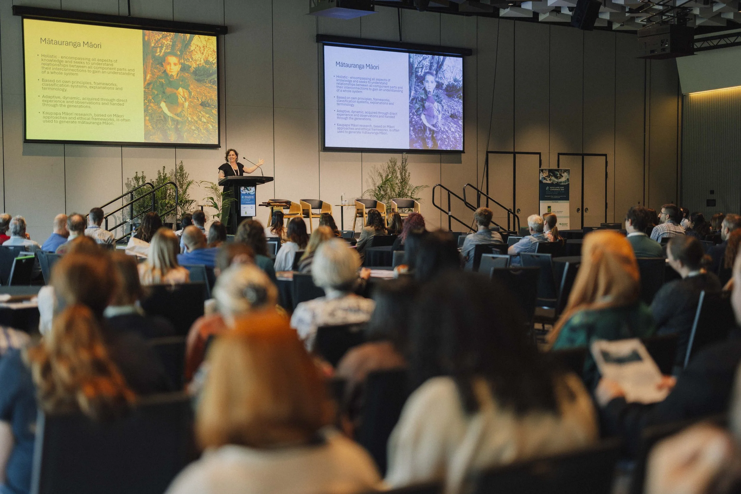 A woman giving a presentation at a conference in a large room filled with seated audience members, with two large screens displaying a slide titled 'Mātauranga Māori' and a photo of a young girl.
