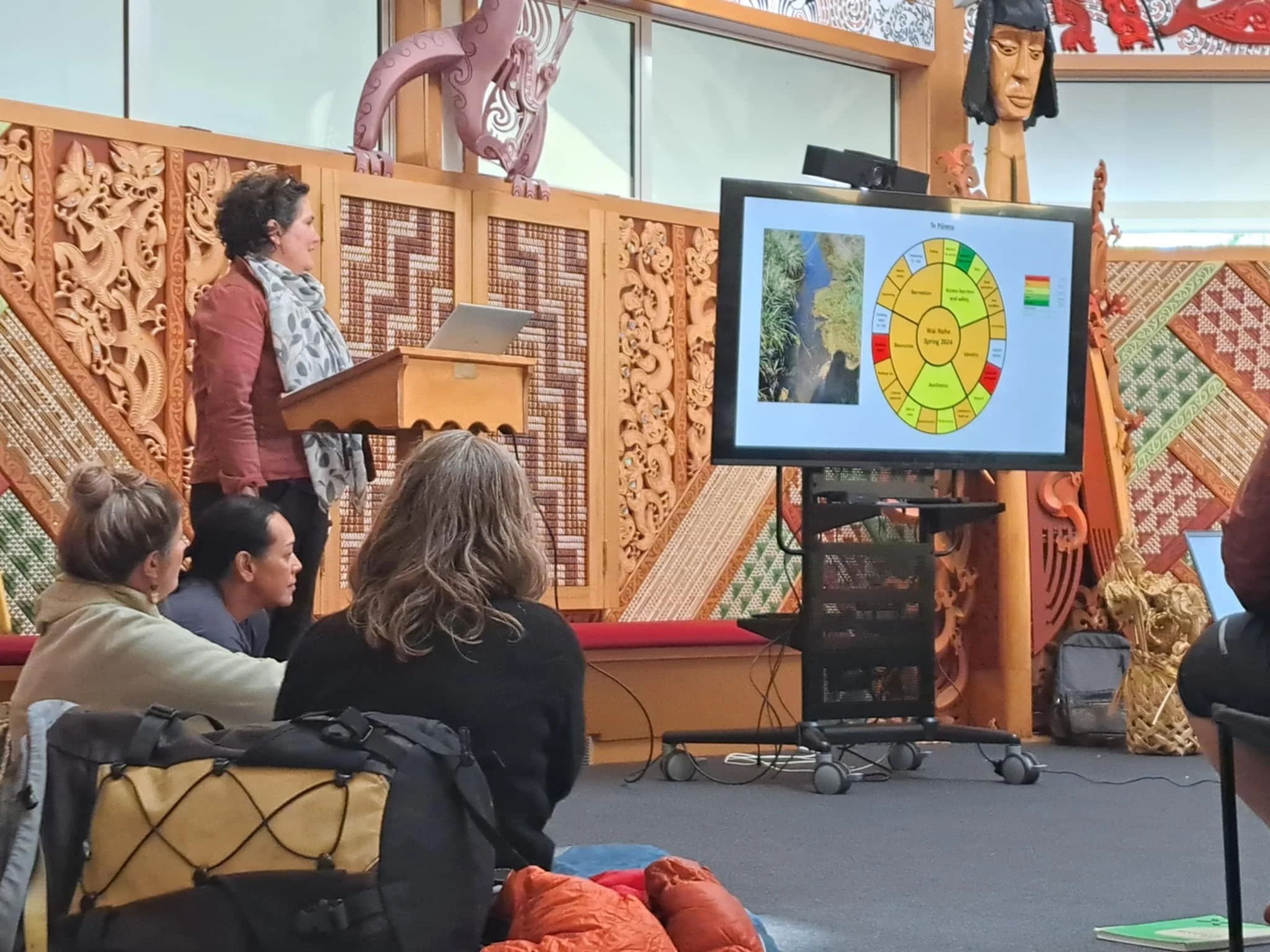 A woman is giving a presentation to an audience in a room decorated with traditional wooden carvings. She stands at a lectern next to a large screen displaying a diagram and images related to the topic. Several people are seated, listening attentively.