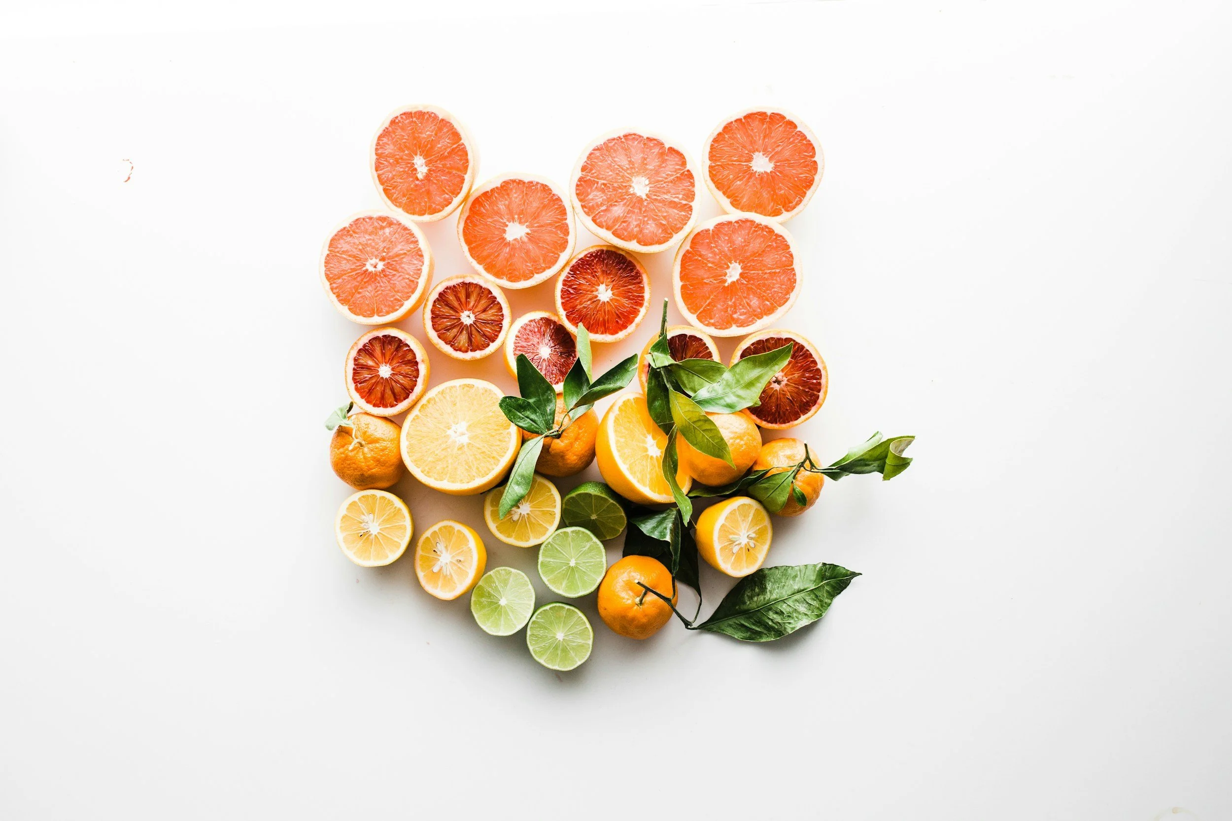 A variety of sliced citrus fruits including oranges, lemons, limes, and blood oranges arranged on a white background with some green leaves.