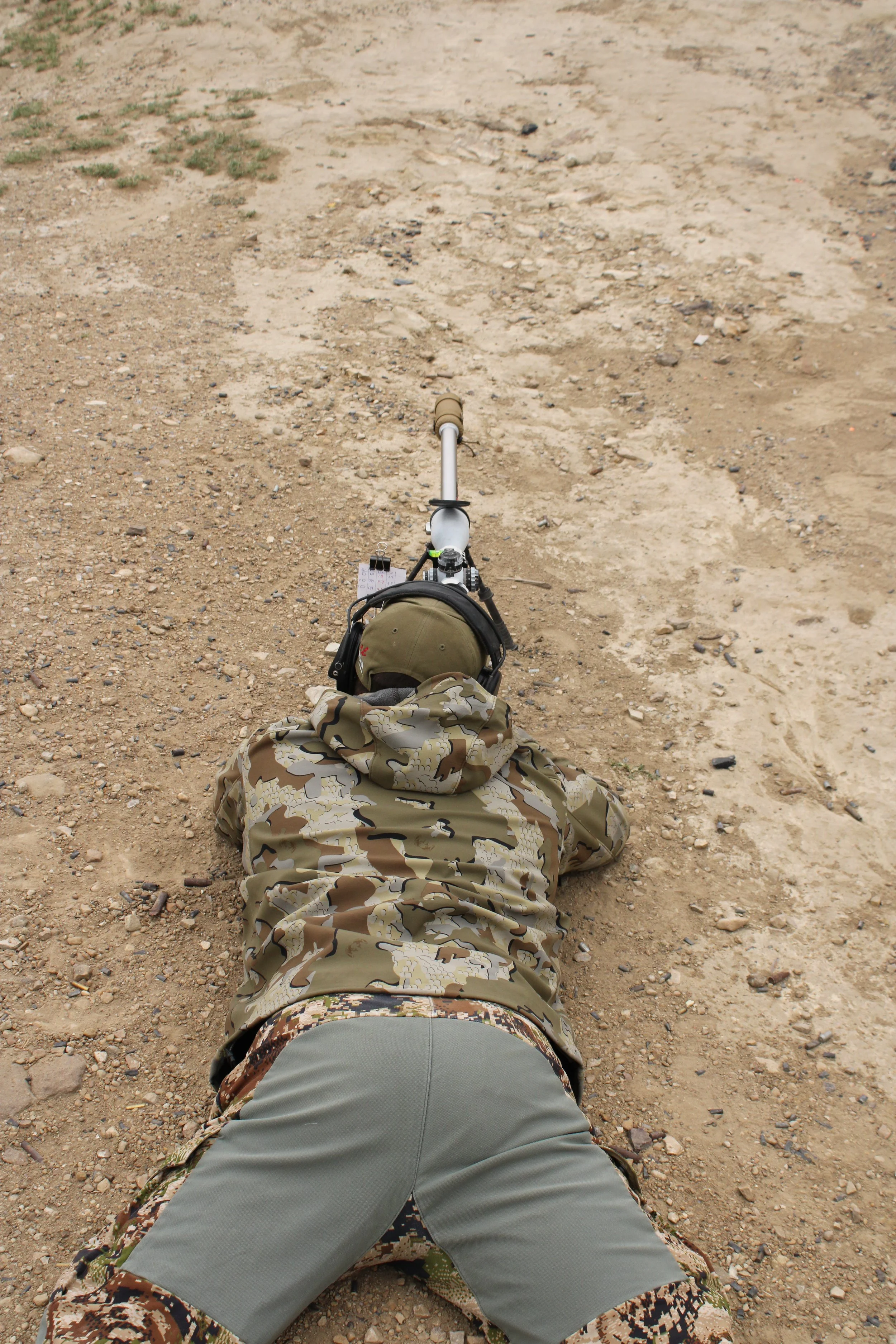 A person lying on the ground in a desert-like area, wearing camouflage clothing and a tan hat, with a drone on the ground nearby.
