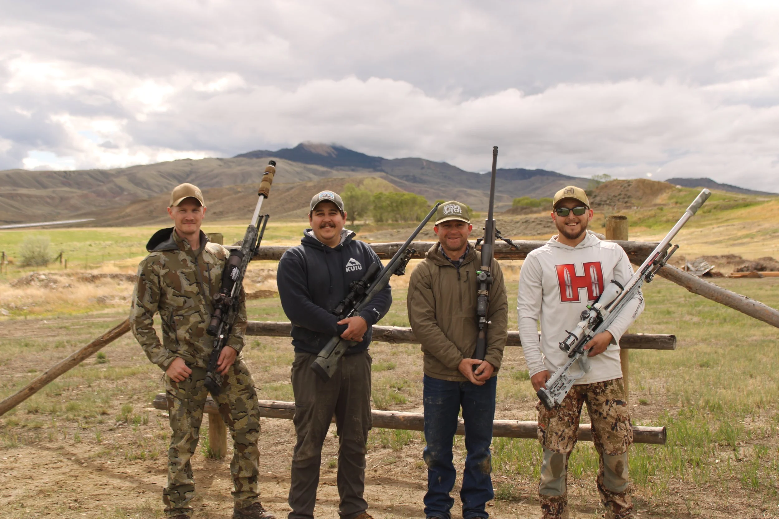 Four men standing outdoors behind a wooden fence, holding rifles, with mountains and cloudy sky in the background.