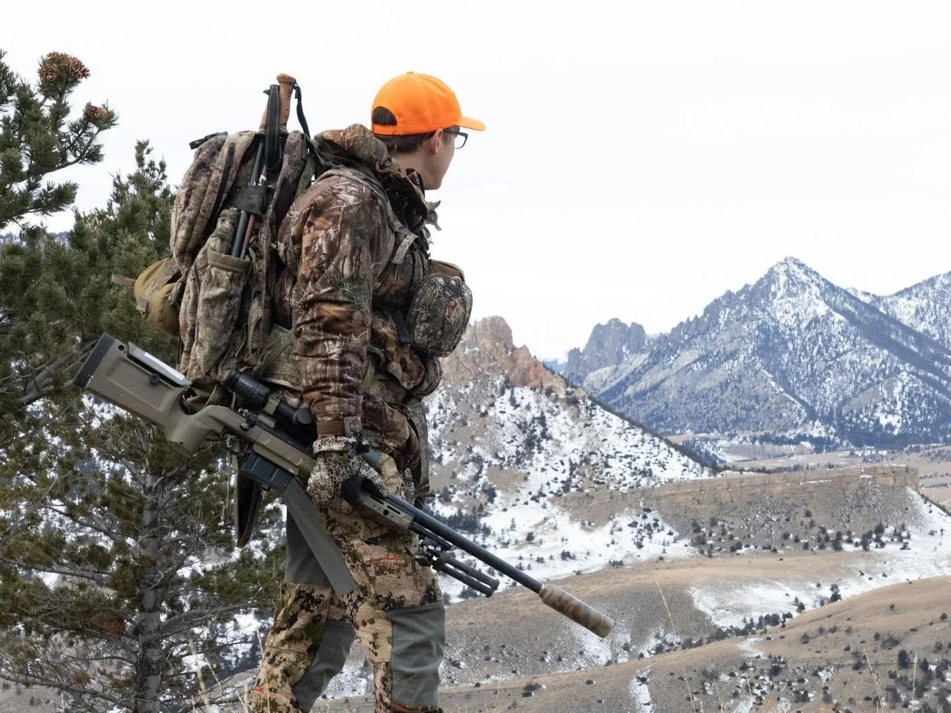 Hunter in camouflage clothing carrying a rifle in a snowy mountain landscape with pine trees and rugged peaks.