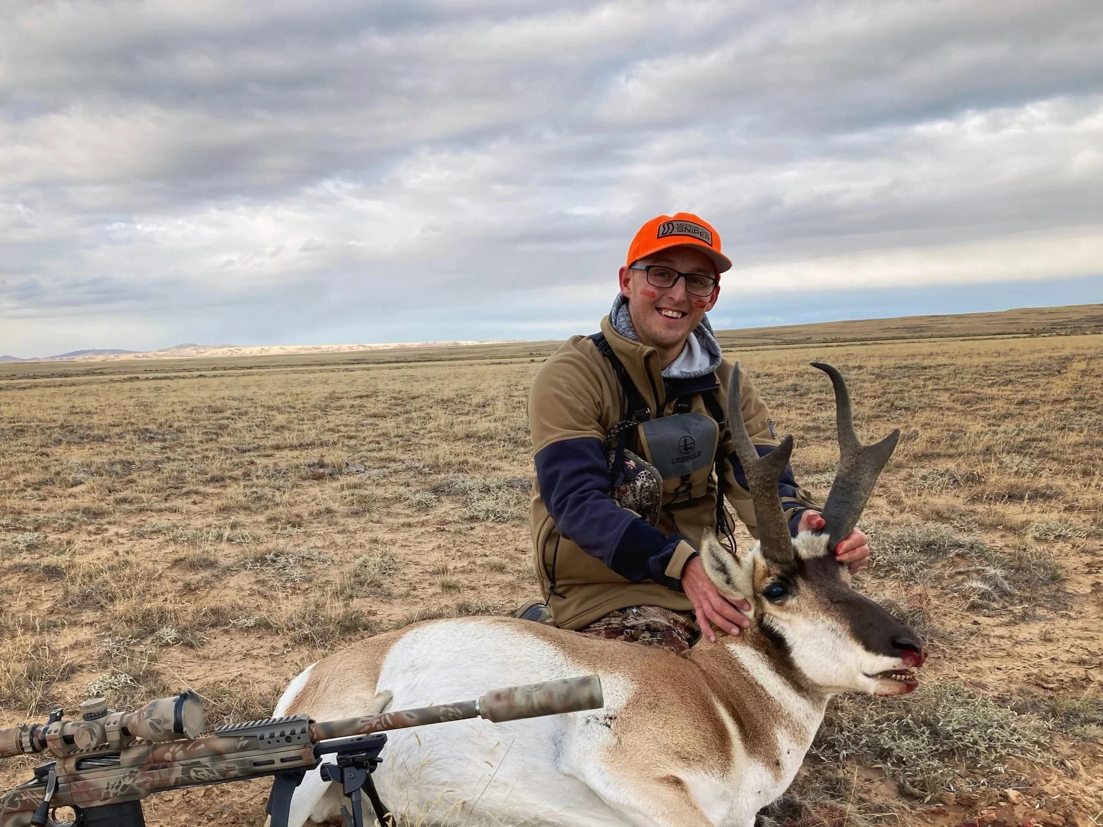 A man in outdoor clothing, wearing glasses and an orange cap, smiling while holding a deer with antlers in a grassy, open landscape under a cloudy sky. A bolt-action rifle with a scope rests on the ground near him.