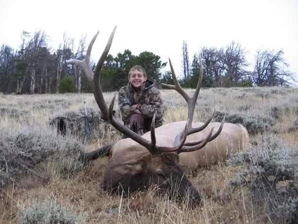 A young boy sitting on a large elk with impressive antlers, in a grassy outdoor setting with trees in the background.