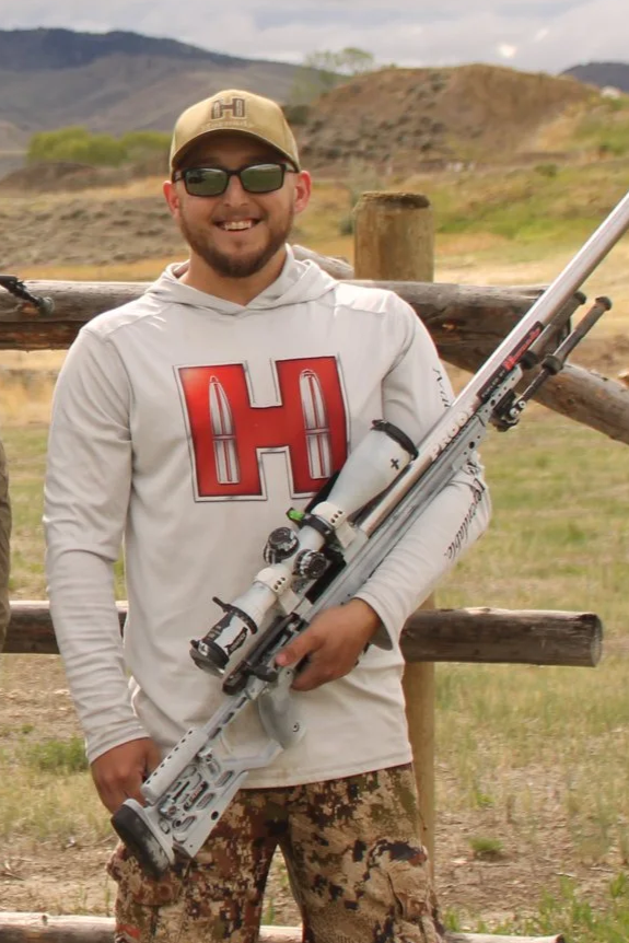 A man standing outdoors holding a sniper rifle, wearing camouflage pants, a light gray hoodie with a large red letter 'H' on it, a beige cap with an 'H' logo, and sunglasses, with a rural landscape in the background.