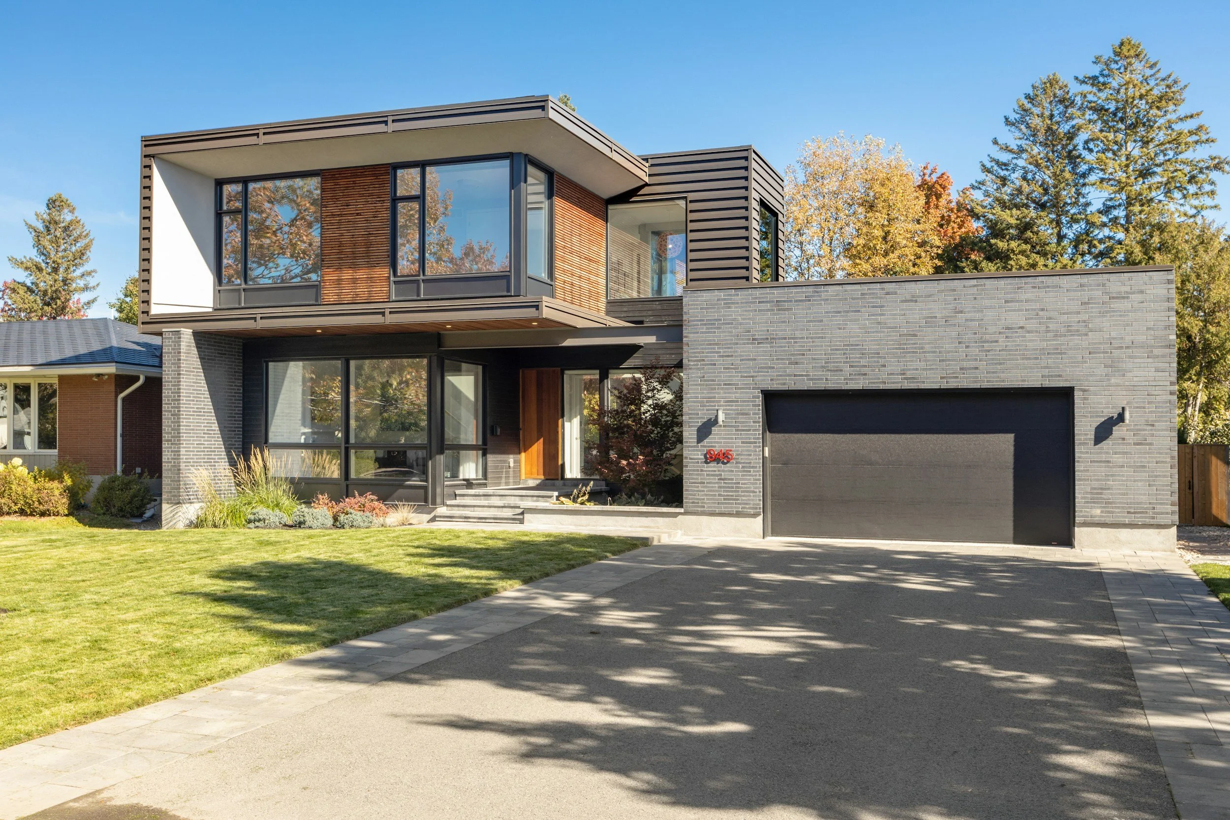 Modern two-story house with gray brick exterior, large glass windows, and a black garage door. Front yard with green grass and concrete driveway, surrounded by trees with autumn foliage, under a clear blue sky.