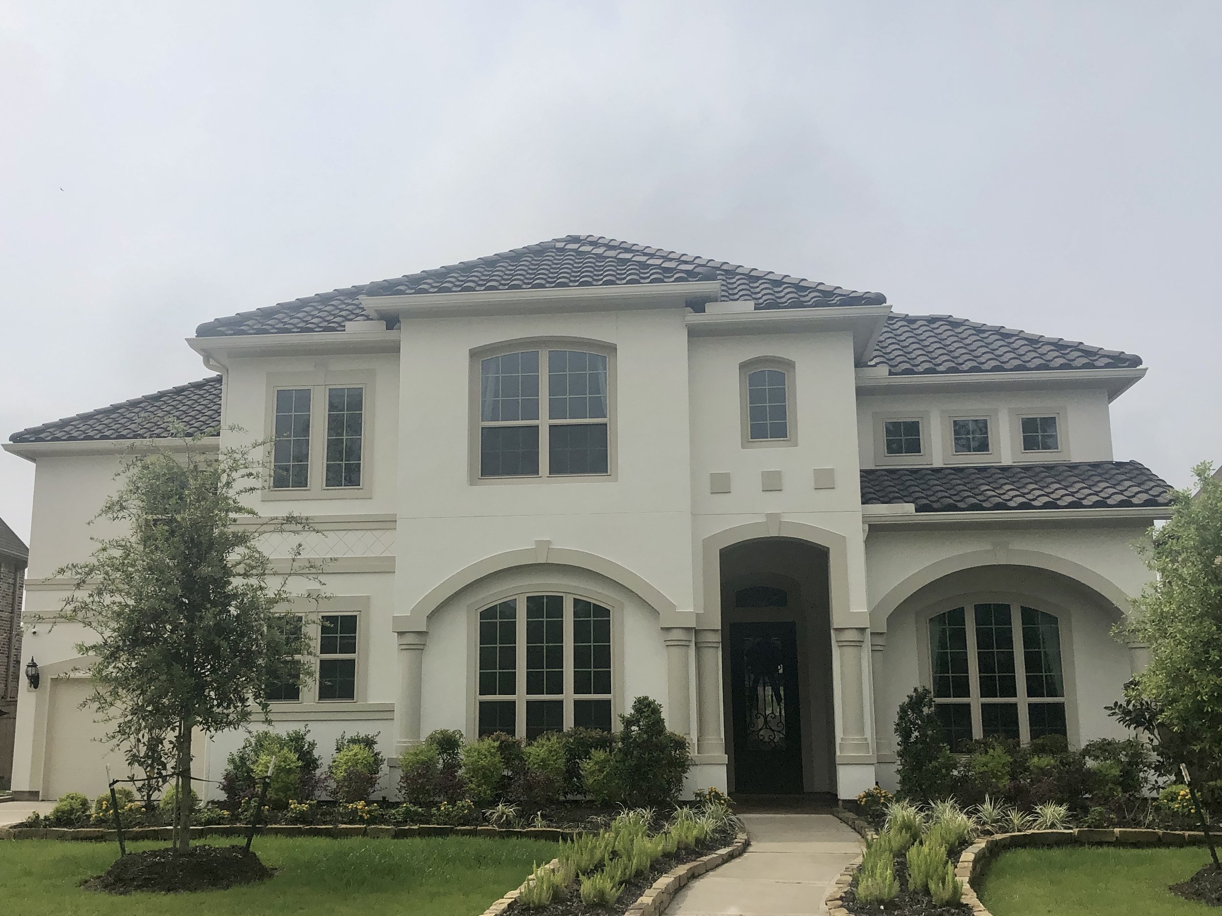 A large white house with multiple windows and a tiled roof, surrounded by a landscaped front yard with a curved walkway, small trees, and green bushes, under an overcast sky.