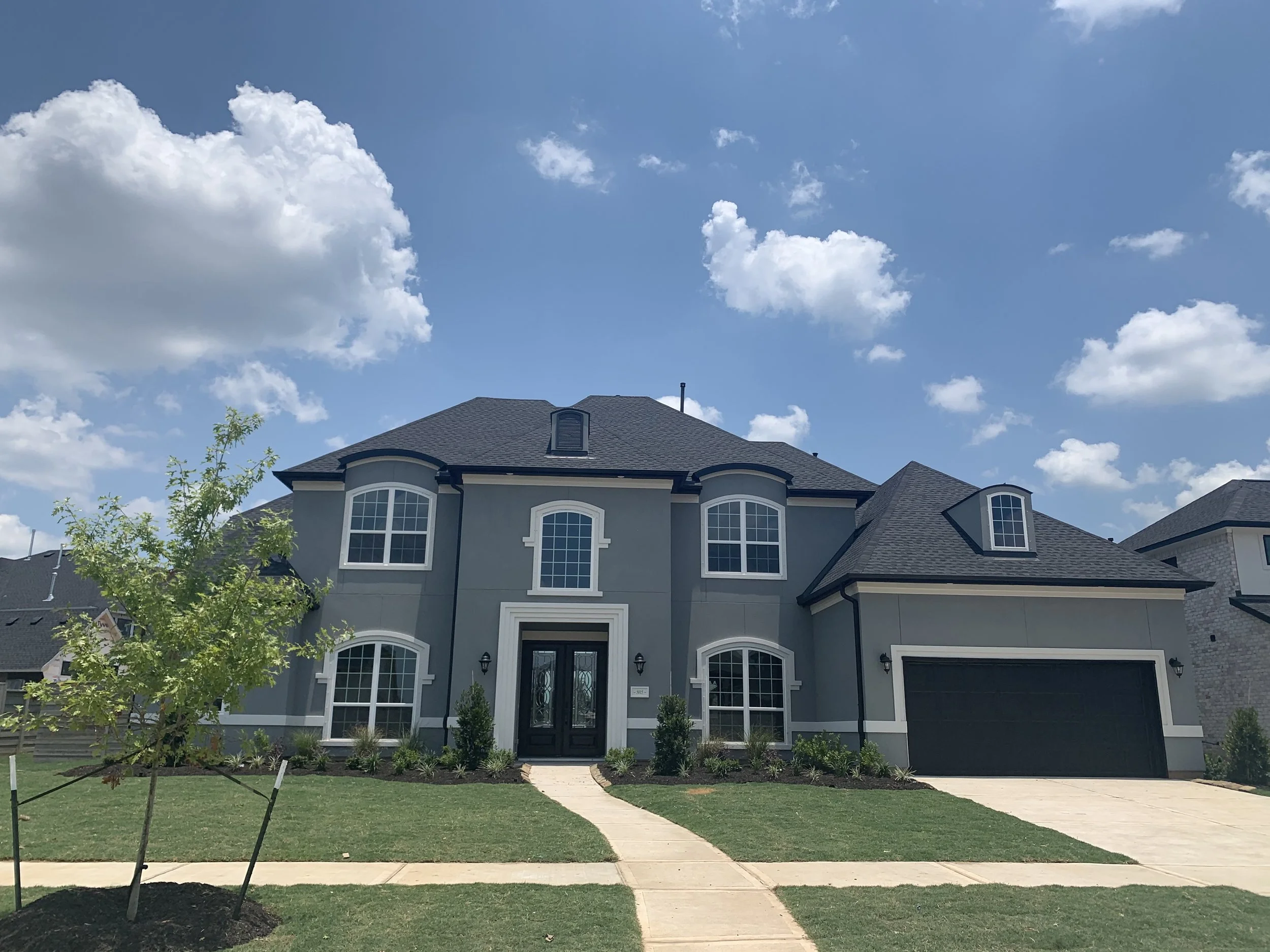 Front view of a two-story house with gray exterior, black door, and white window frames, surrounded by a green lawn, small tree, and a clear blue sky with white clouds.