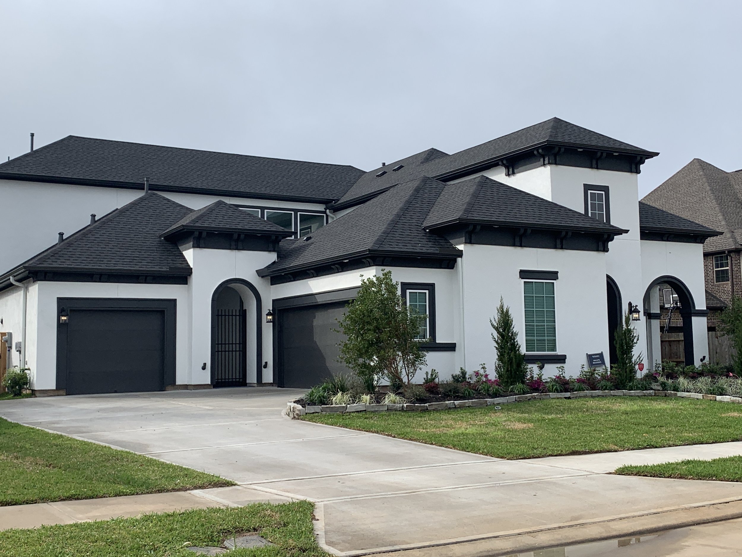 A modern townhouse with a white exterior, black accents, and multiple peaked shingle roofs. There is a driveway, a small landscaped yard with bushes and flowers, and a cloudy sky above.
