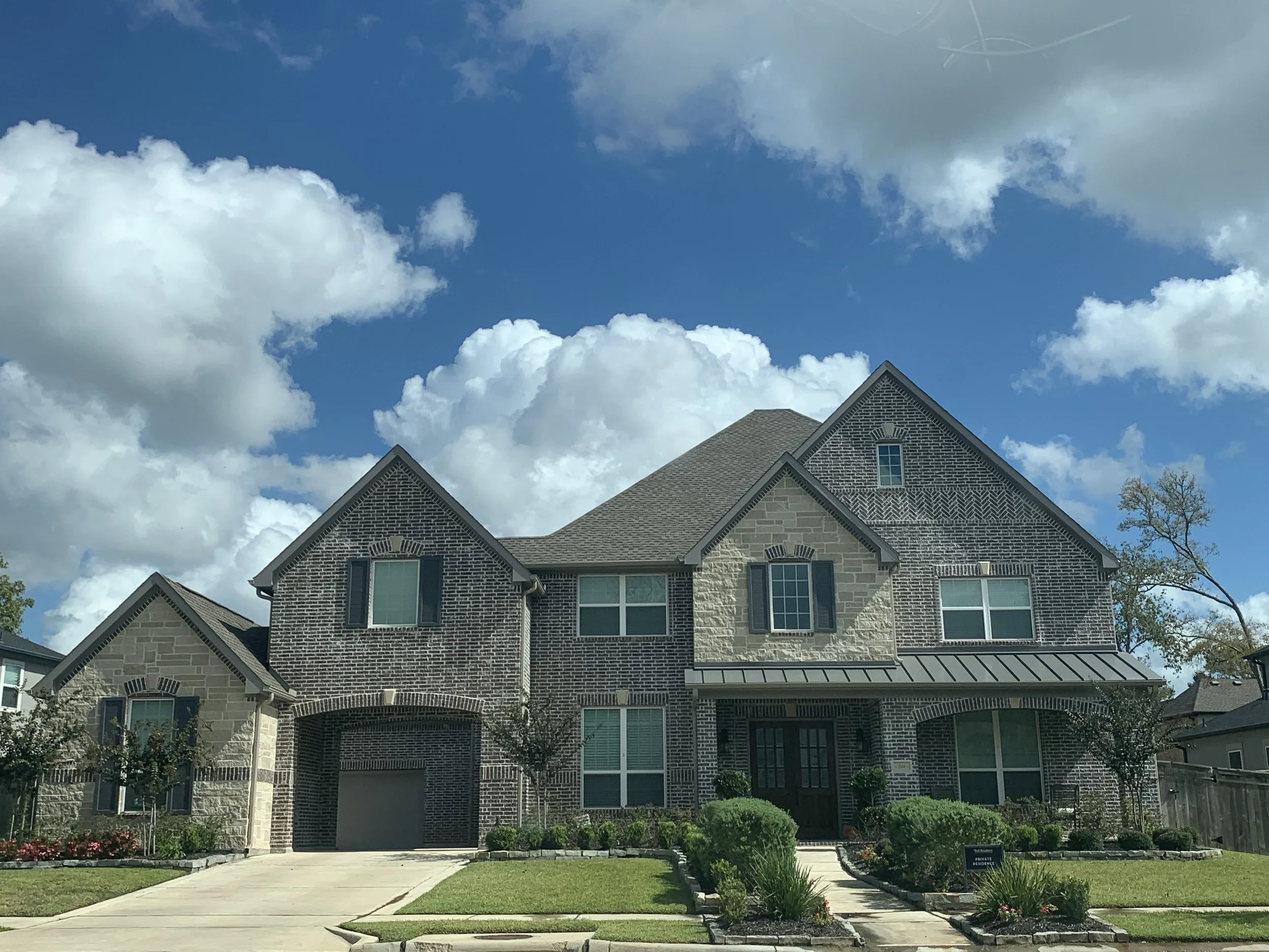 A modern brick and stone house with multiple gable roofs, large windows, a garage door, and a front garden with green grass, small bushes, and flowering plants. The sky above is bright blue with puffy white clouds.