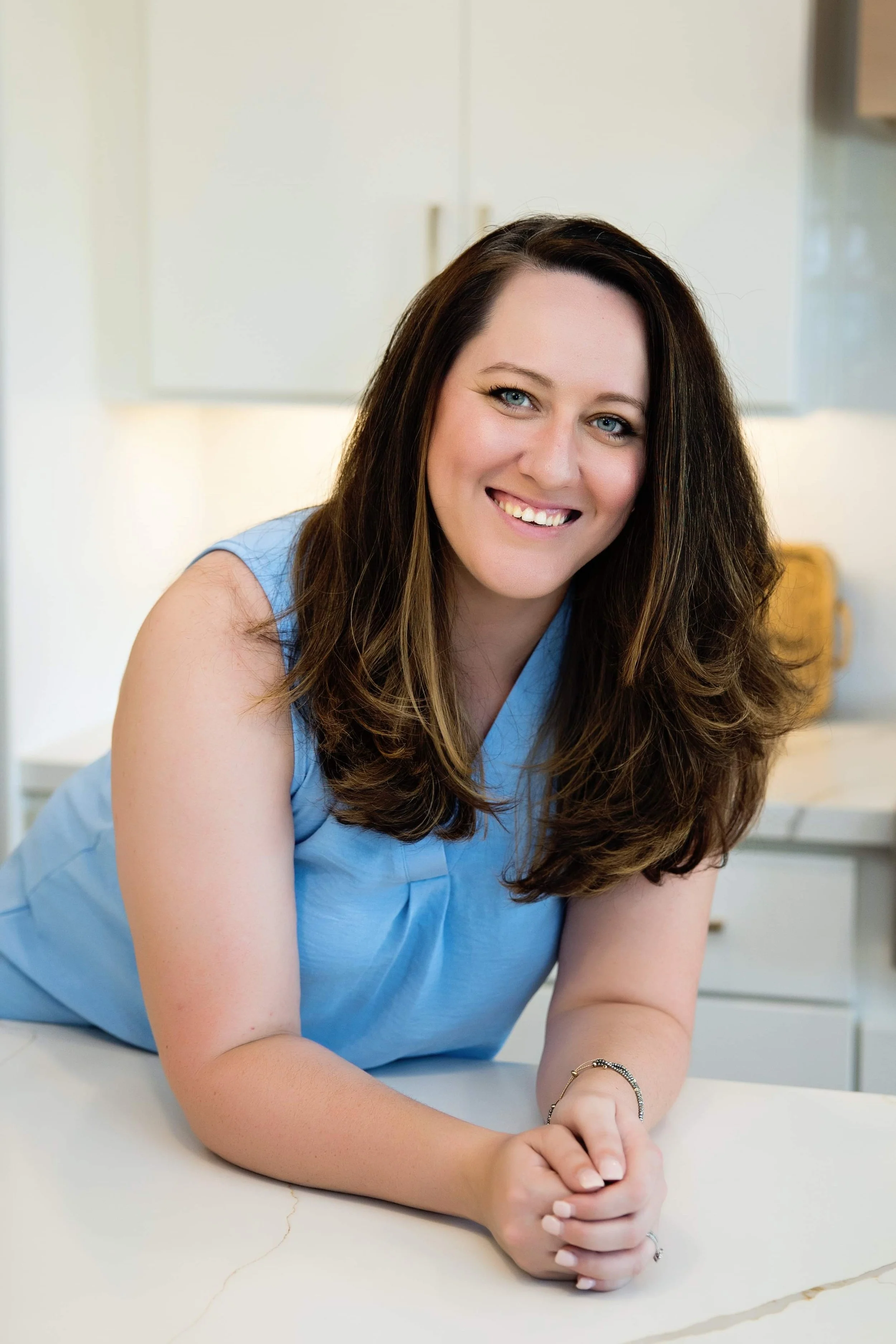A woman with long brown hair and blue eyes, smiling while leaning on a white table in a bright kitchen, wearing a sleeveless blue top.
