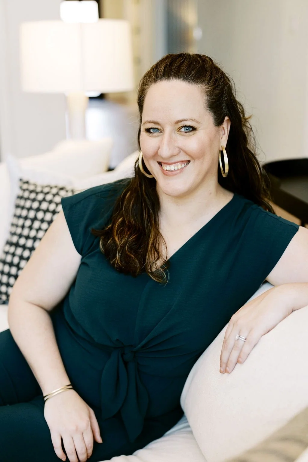 A woman with long dark hair, wearing a teal top and gold hoop earrings, sits on a light-colored sofa, smiling at the camera in a well-lit living room.