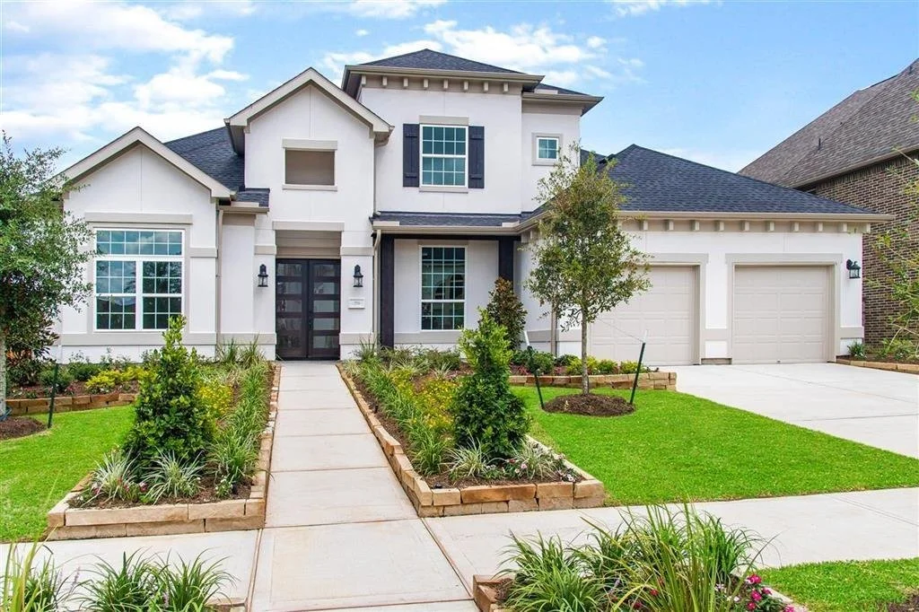 Front view of a modern two-story house with a white exterior and dark roof, landscaped front yard with shrubs, flowers, and small trees, concrete walkway leading to the entrance, and a two-car garage with white doors.