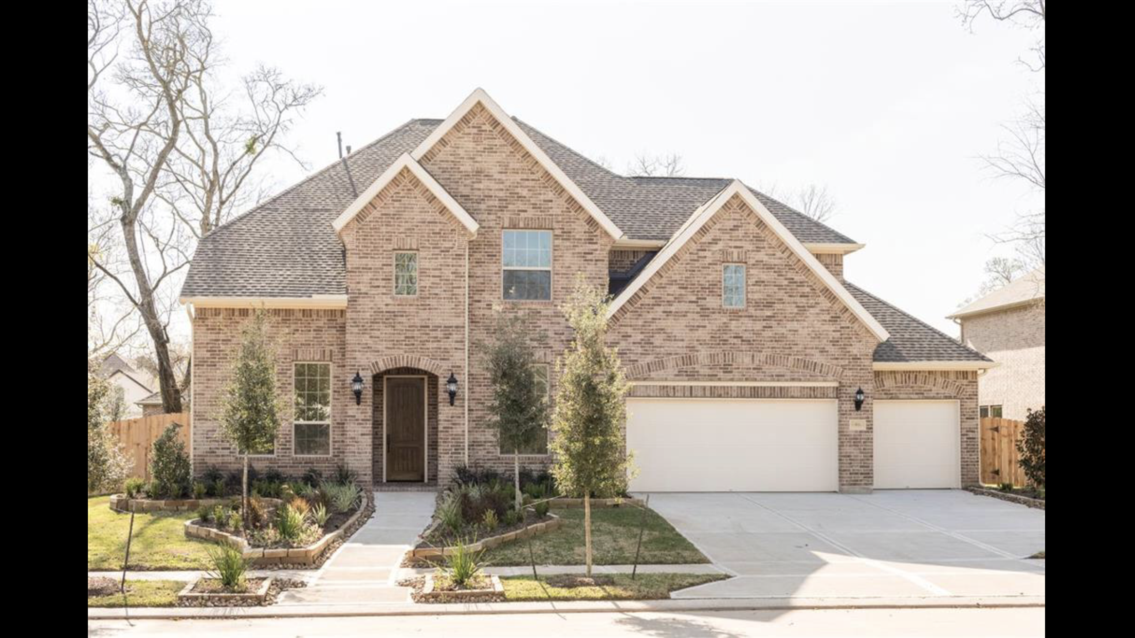 Front view of a two-story brick house with a brown front door, two garage doors, and a landscaped yard with small trees and shrubs.