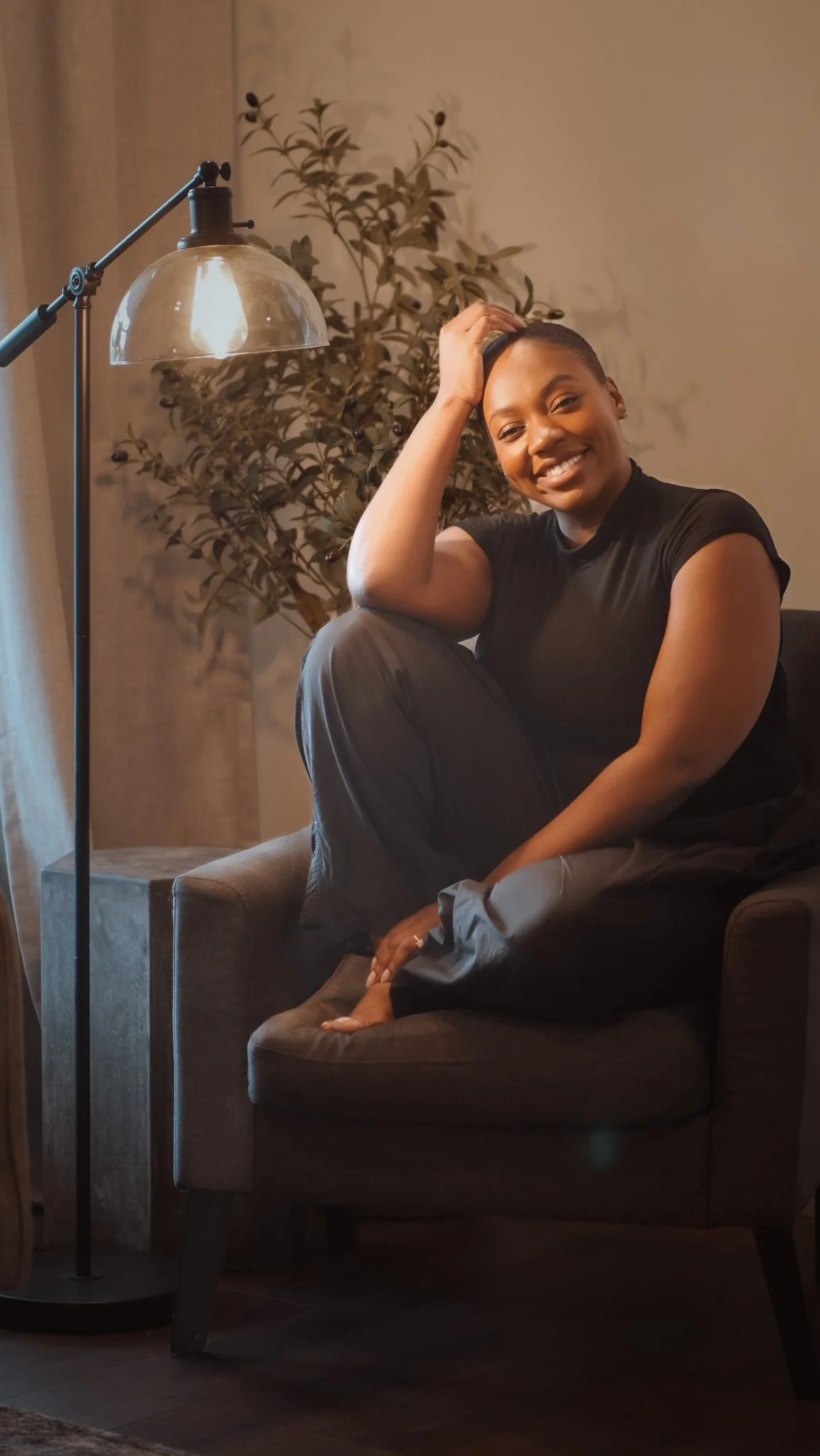 A smiling woman sitting on a brown armchair, with her right hand in hair. She is wearing a black t-shirt and gray pants. There is a tall floor lamp with a metal shade to her left and a large green plant in the background. The setting appears to be indoors with warm lighting.