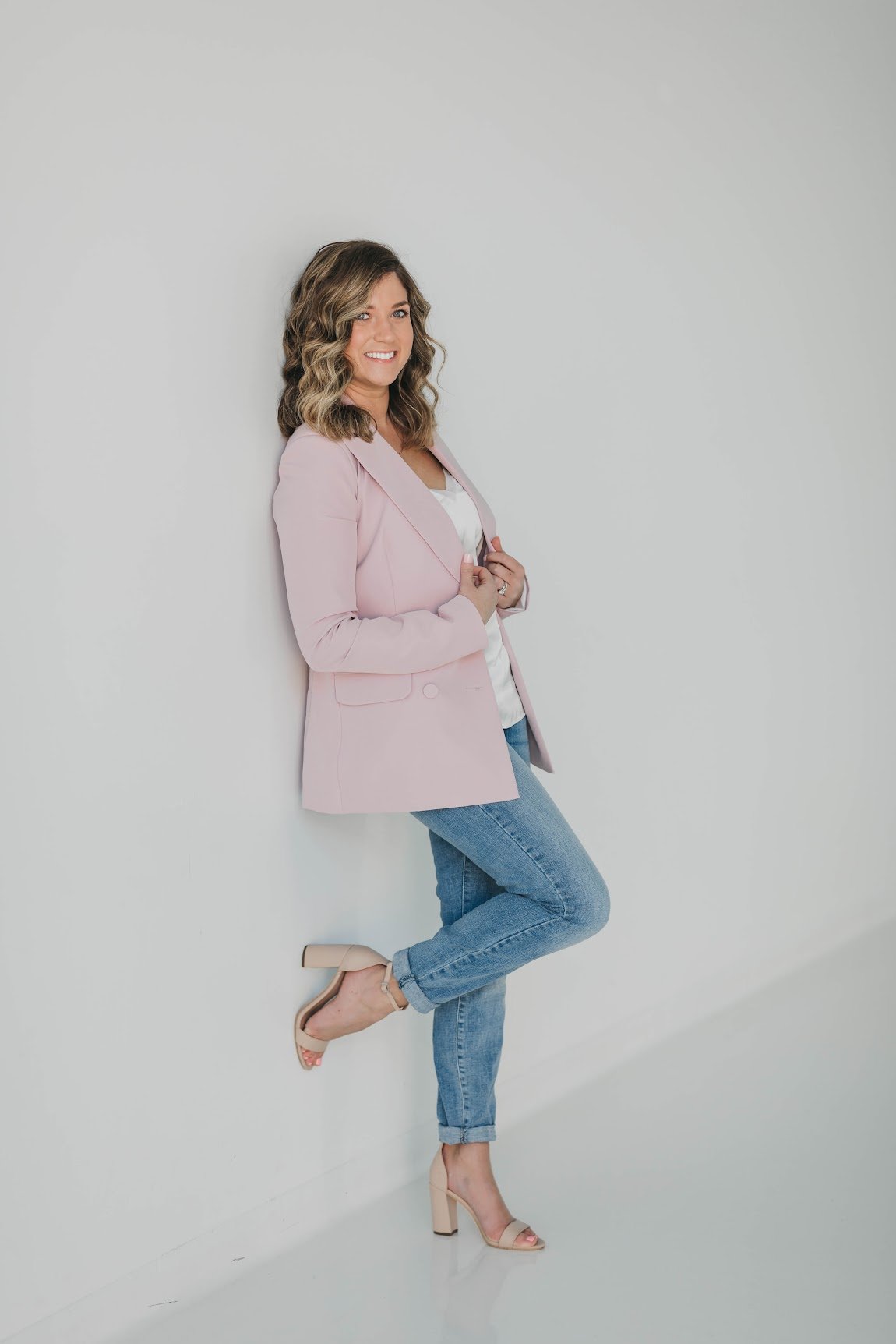Woman in light pink blazer and blue jeans leaning against white wall, smiling, with one foot raised and beige high heels.