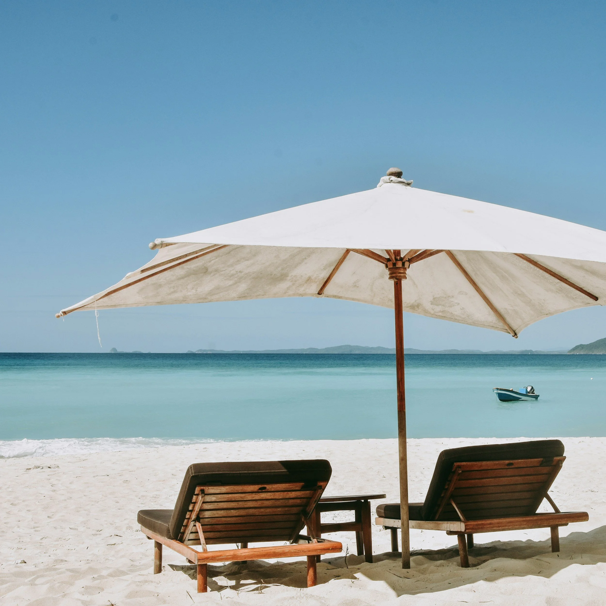 Beach scene with two wooden lounge chairs and a large white beach umbrella, overlooking the ocean with a boat in the water and mountain in the background under a clear blue sky.
