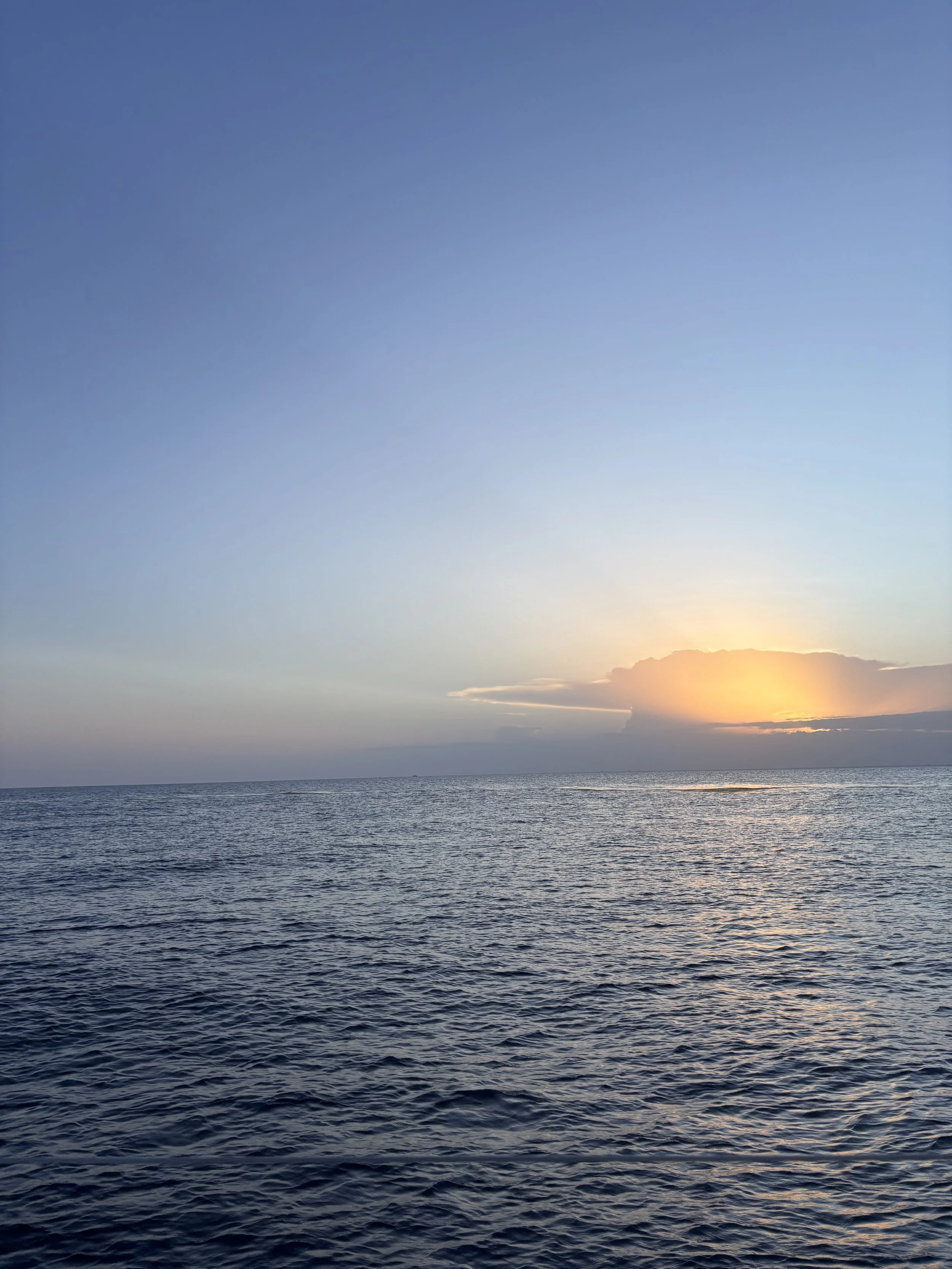 A calm ocean with gentle waves at sunset, with a partially clouded sky and the sun near the horizon.