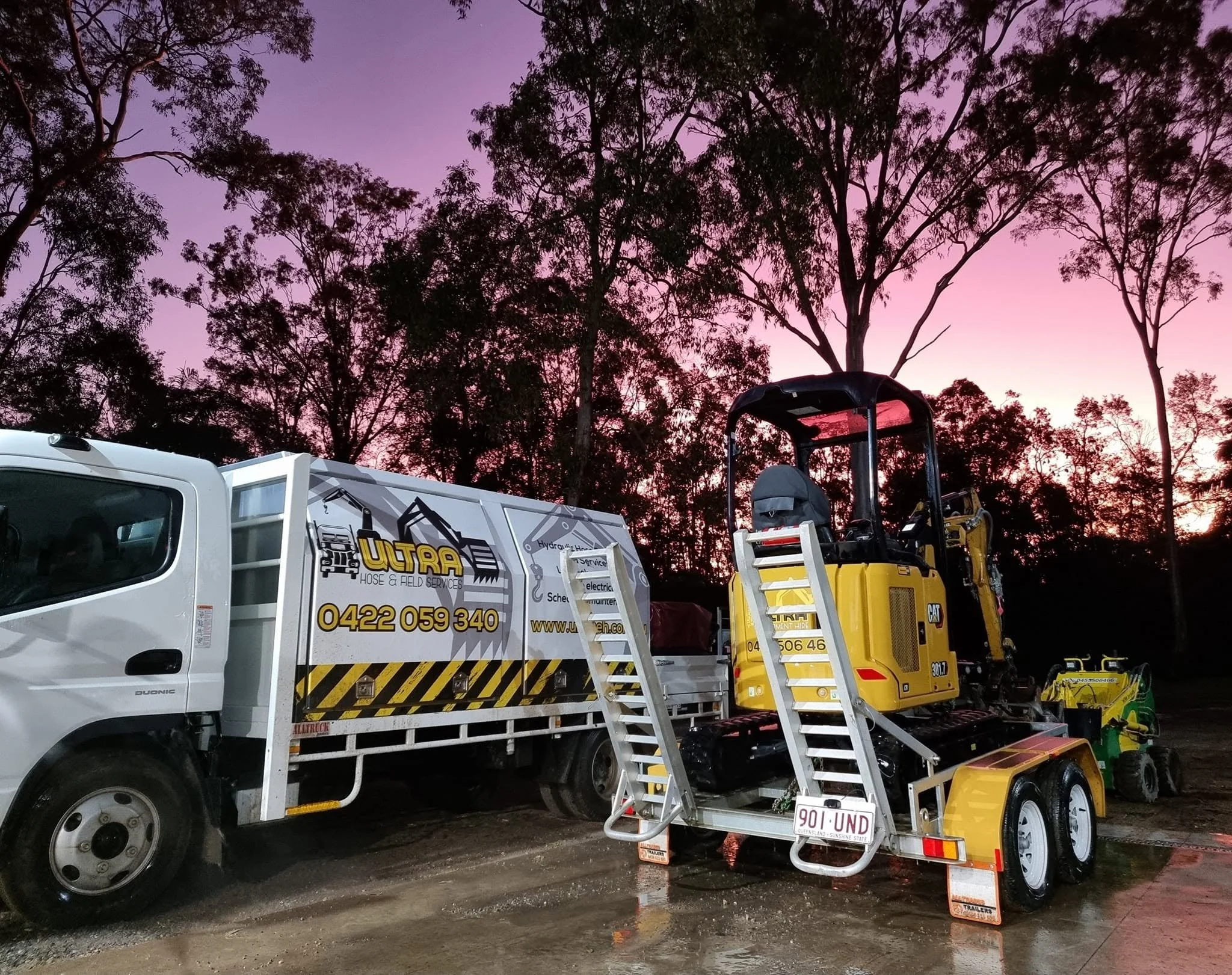 Construction vehicles and equipment parked outdoors at sunset with purple and pink sky, including a white utility truck, a yellow construction machine, and various tools and supplies.