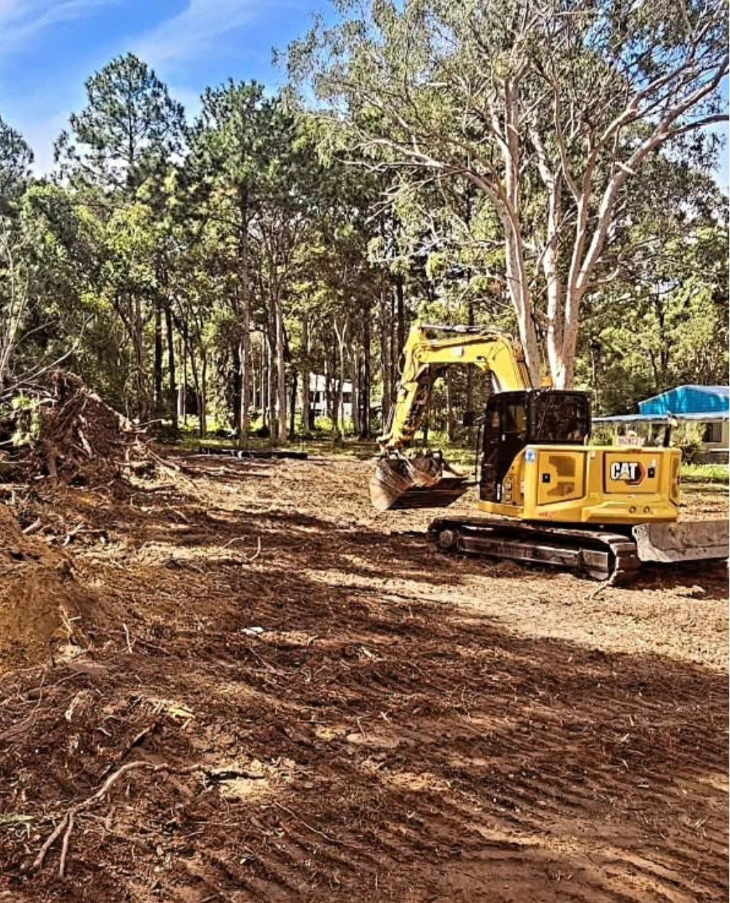 A yellow excavator on a dirt area clearing land with trees in the background and a blue sky overhead.