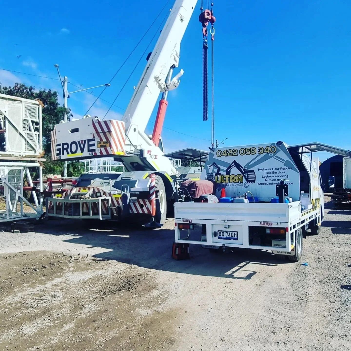 A construction site with a white crane truck labeled 'GROVE' and a white work truck with contact information for ULTRA FIELD SERVICES, along with construction equipment and materials on a dirt surface under a clear blue sky.