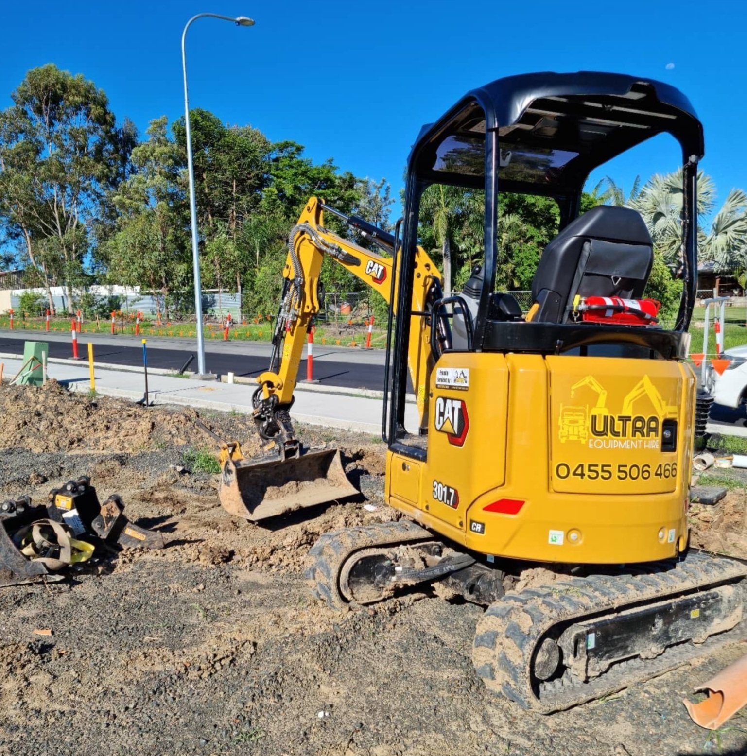 A small yellow CAT compact excavator working on a construction site with dirt and soil, surrounded by trees and clear blue sky.