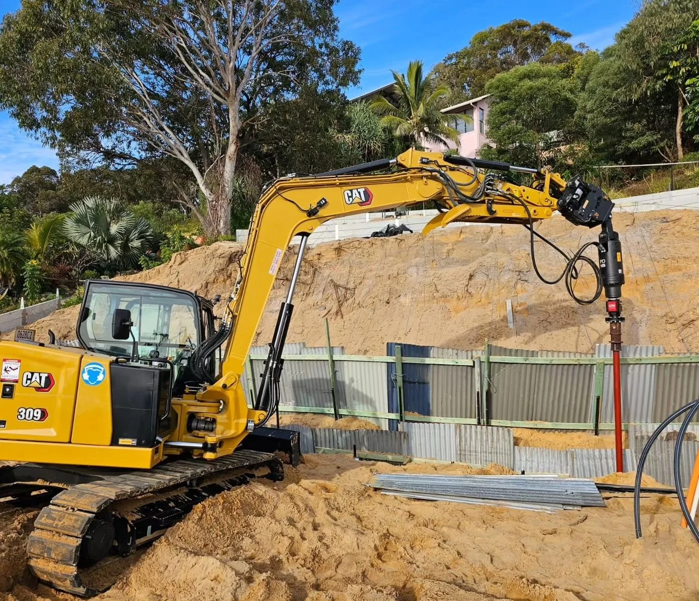 A yellow Caterpillar excavator at a construction site with sandy terrain, a metal fence, and trees and houses in the background.