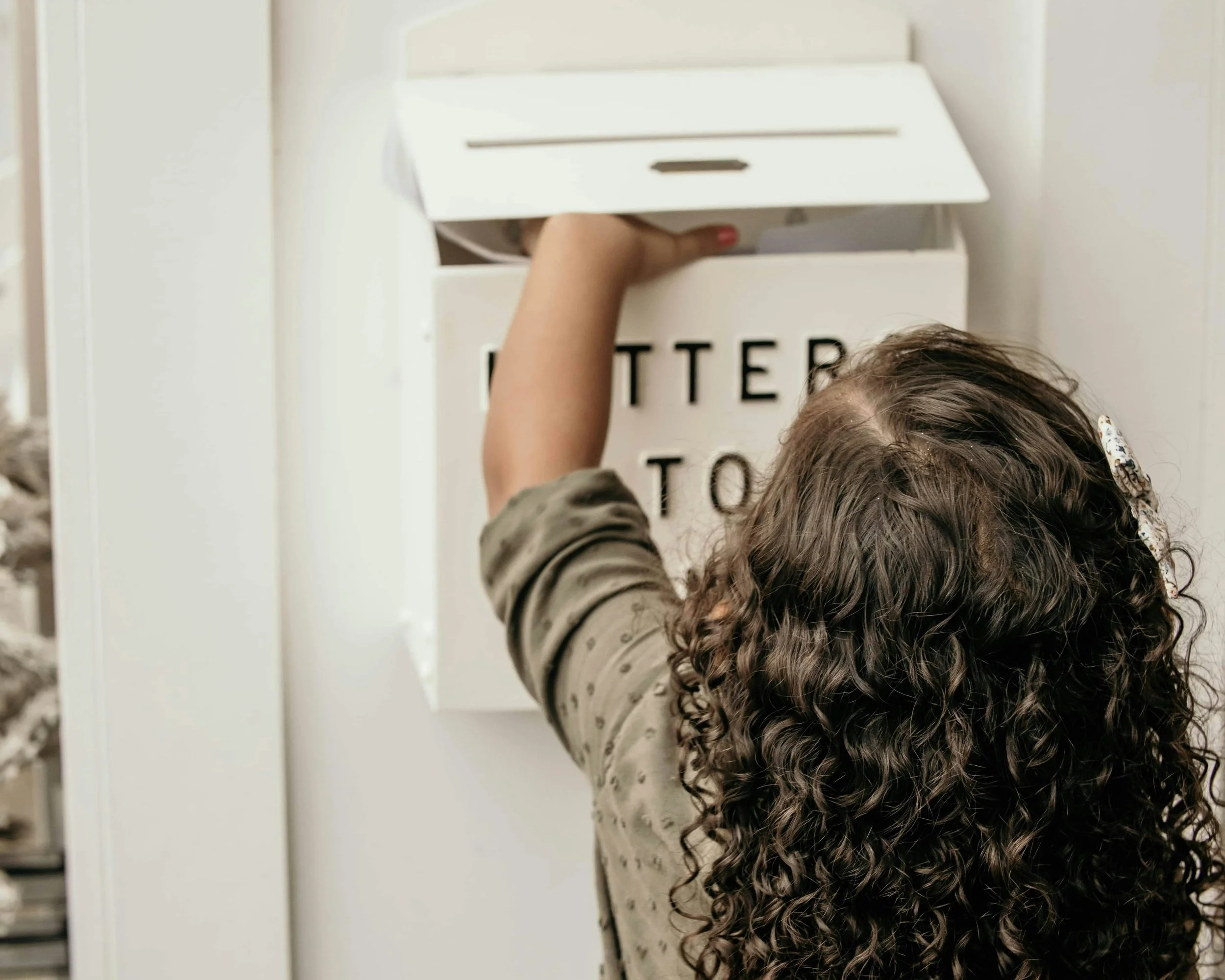 A child with curly hair reaching into a white mailbox.