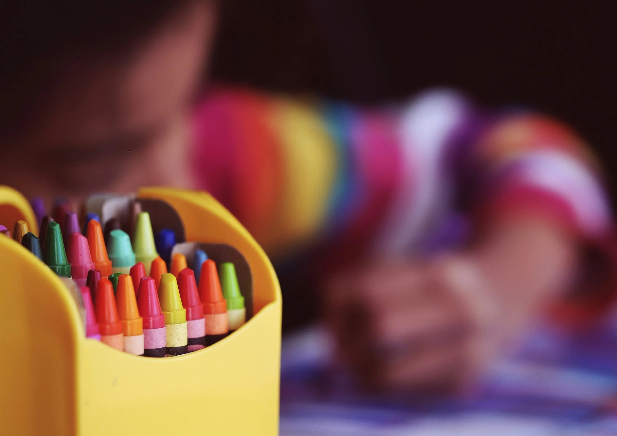 Close-up of a yellow crayon box containing colorful crayons, with a child coloring blurred in the background.