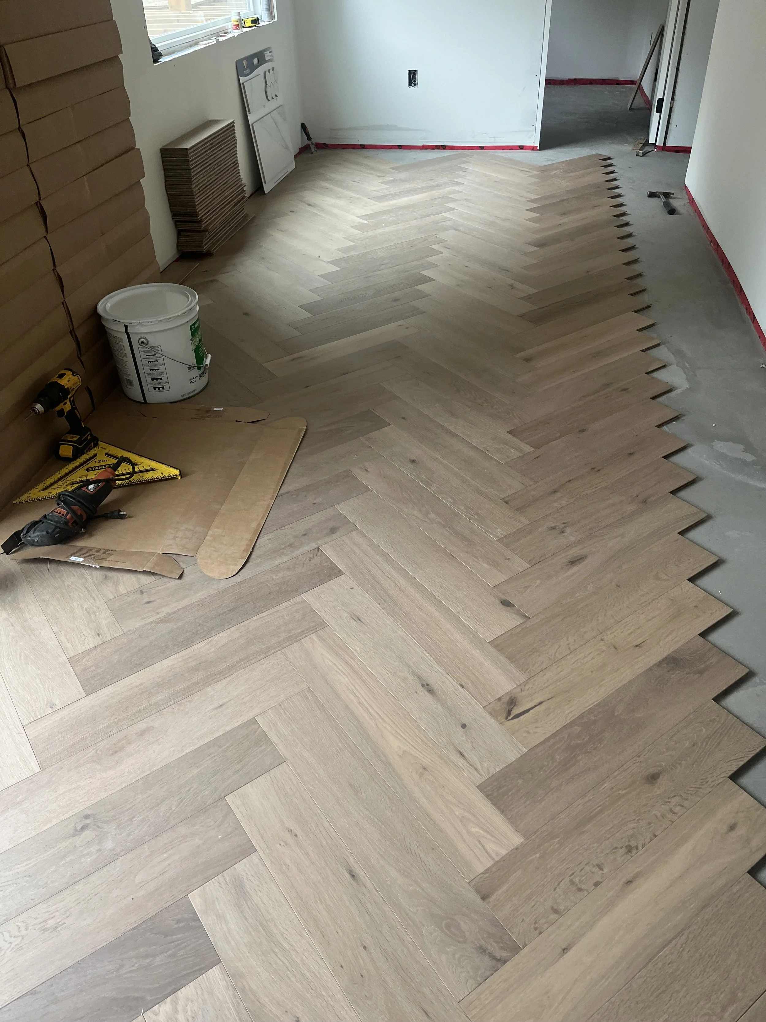 Inside a room under construction, new wooden floor tiles are being installed in a herringbone pattern. Tools and materials such as a drill, saw, and paint bucket are visible on the floor. Walls are unfinished, with some areas taped and prepped for further work.