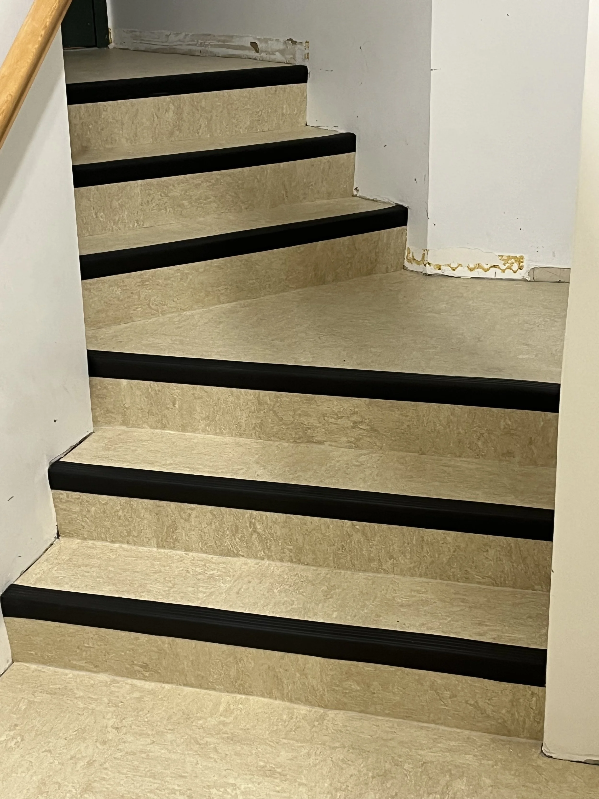 Close-up of a staircase with beige carpeted steps, black anti-slip strips, and a white wall with some peeling paint at the bottom.