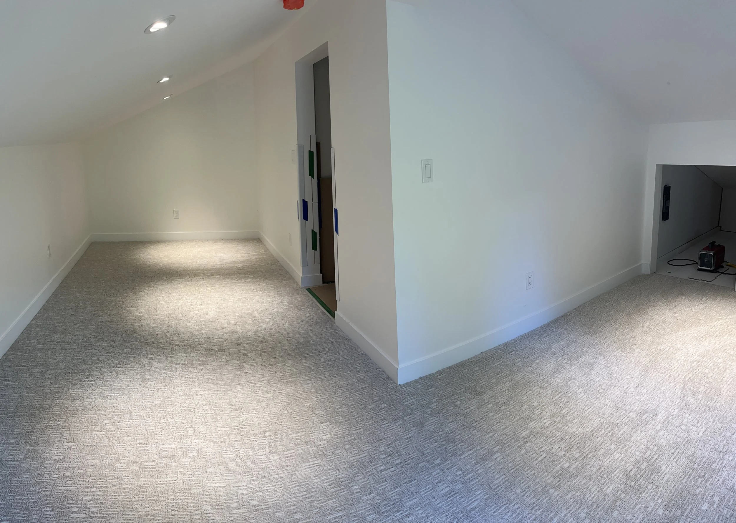 Empty room with beige carpet, white walls, and a sloped ceiling. Recessed ceiling lights and electrical outlets visible.