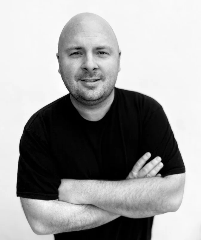 Black and white portrait of a bald man with a beard, smiling, wearing a black t-shirt, standing with arms crossed against a plain background.