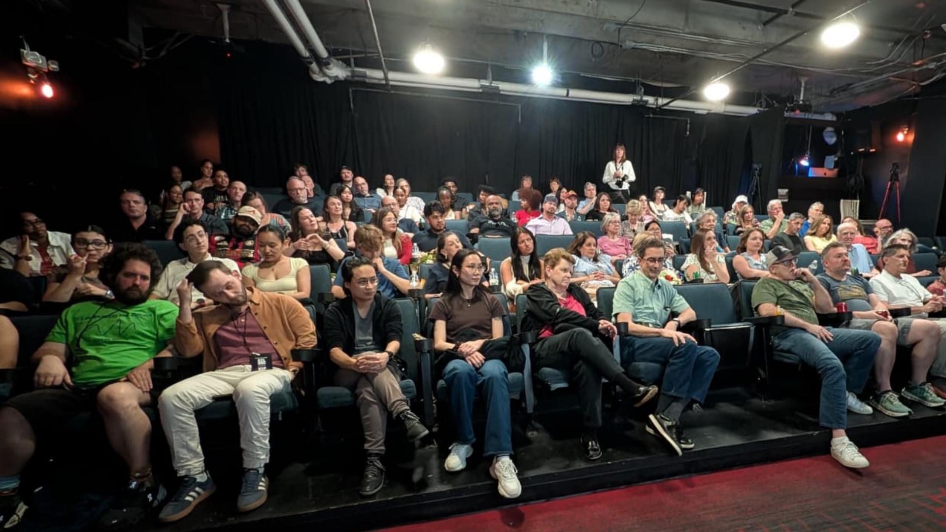 Audience seated in a theater with a dark background, a woman standing on stage, some audience members wearing casual attire, and the room lit with bright overhead lights.