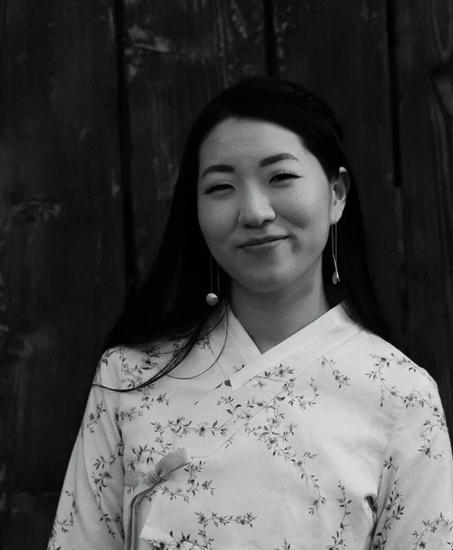 A young woman with long dark hair, smiling slightly with closed lips, wearing a traditional kimono with floral patterns and long earrings, standing in front of a dark wooden background.