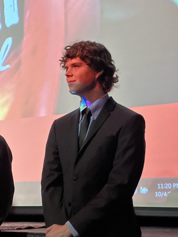 A young man with curly brown hair wearing a dark suit, light-colored dress shirt, and tie, standing in front of a projection screen.