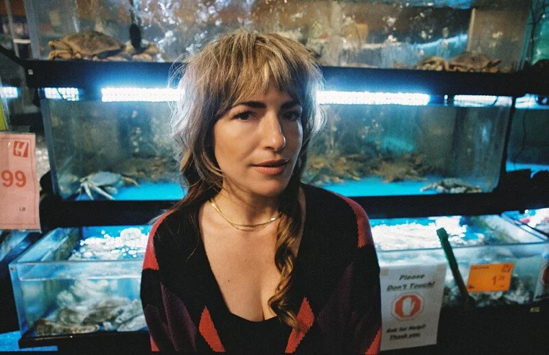 A woman with light brown, wavy hair and layered bangs is standing in front of illuminated aquariums with marine animals inside, in what appears to be an aquarium or pet store.