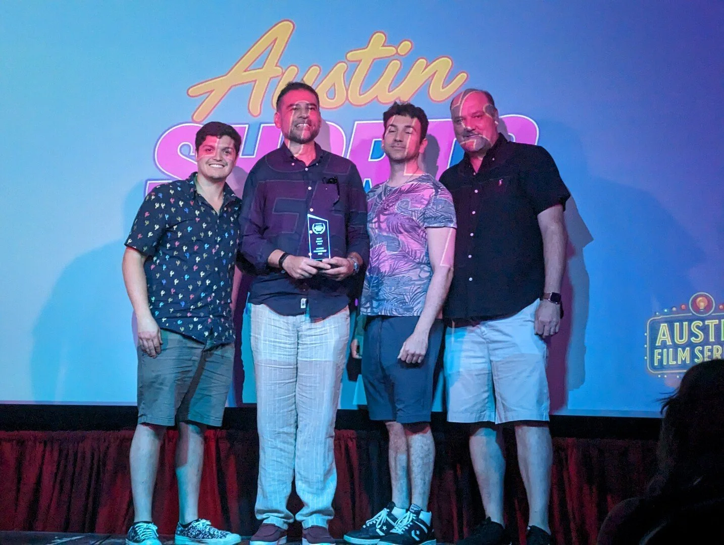 Four men standing on a stage in front of a screen that displays the text 'Austin Shorts' and a logo that reads 'Austin Film Series'. One of the men is holding a trophy or award. They are smiling and dressed casually.